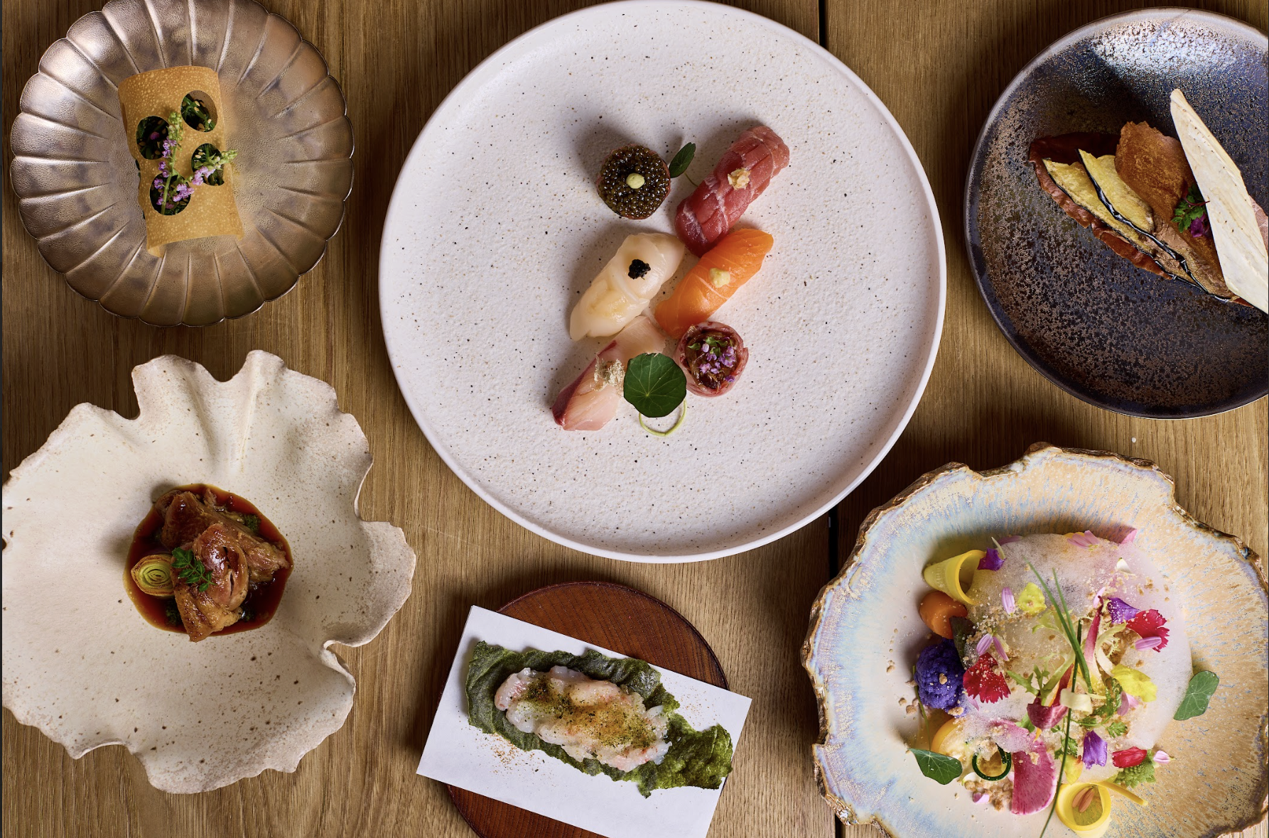 A variety of plated gourmet dishes with colorful garnishes on a wooden table, including small portions of seafood, meat, and vegetable dishes, served on decorative plates.