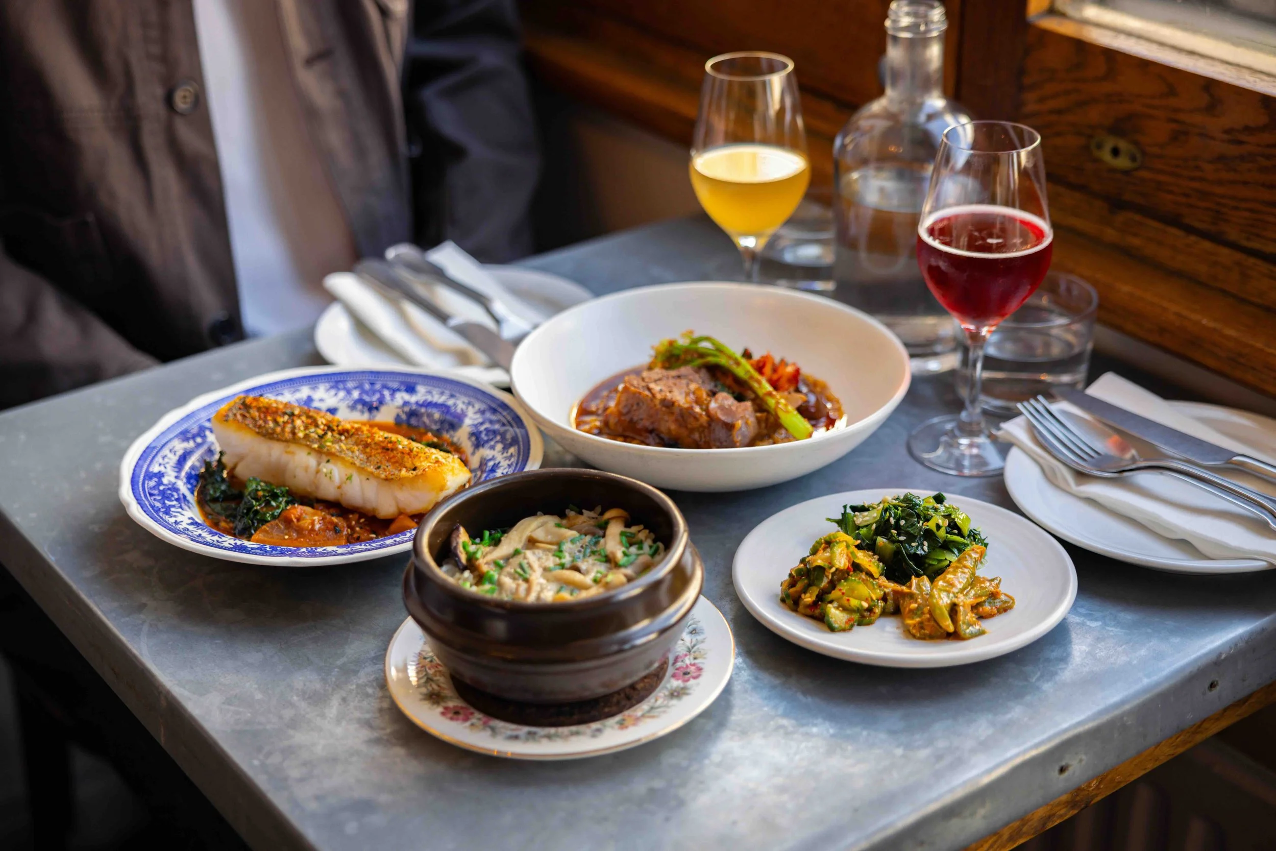 Assorted dishes and beverages on a restaurant table, including a fish fillet with sauce, a bowl of meat stew with vegetables, various side dishes, and glasses of white, red, and sparkling drinks.