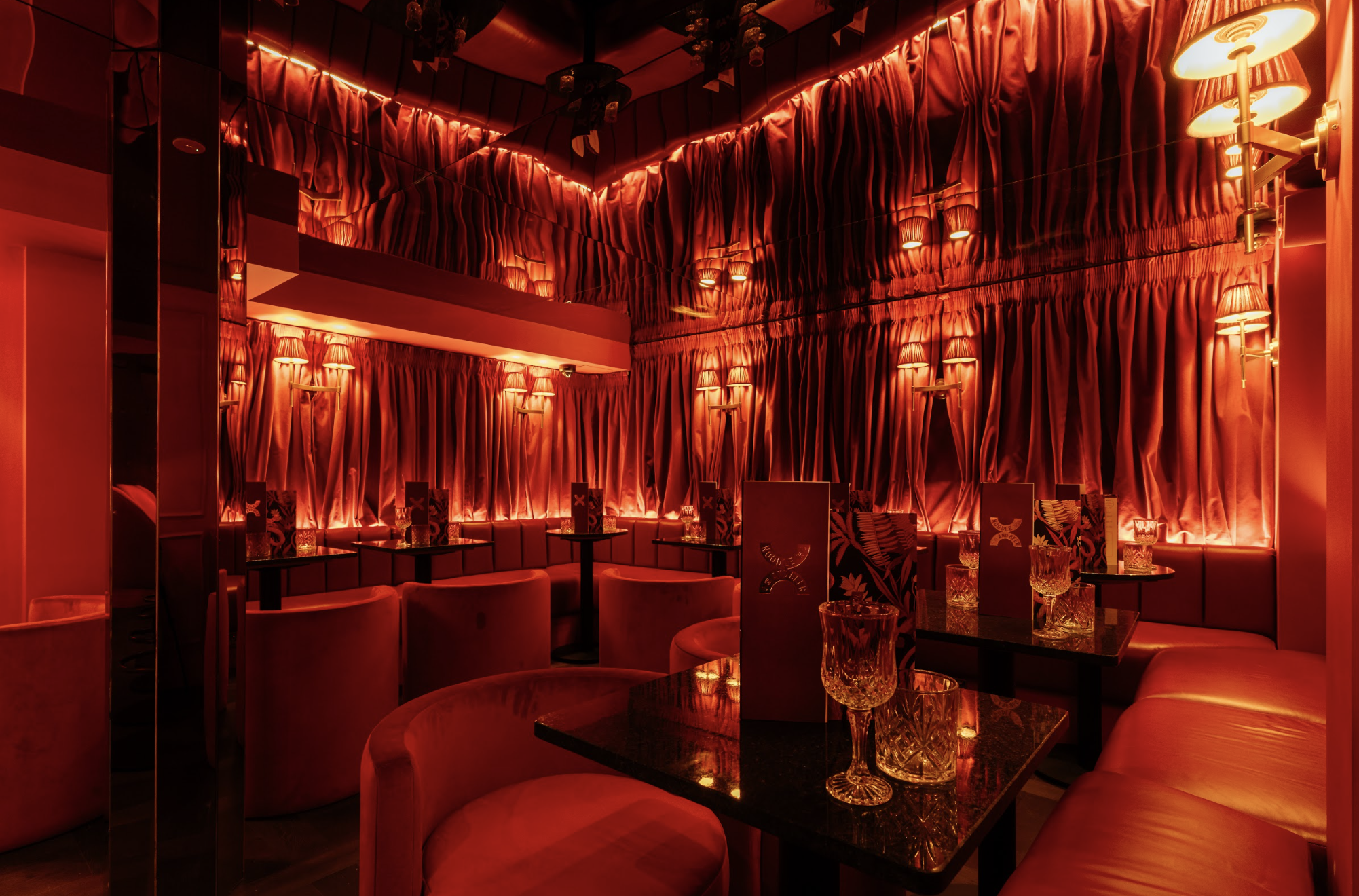 A dimly lit room with red curtains covering the walls and ceiling, illuminated by warm wall-mounted lamps. There are tables with glassware and chairs arranged for dining, creating an intimate, cozy atmosphere.