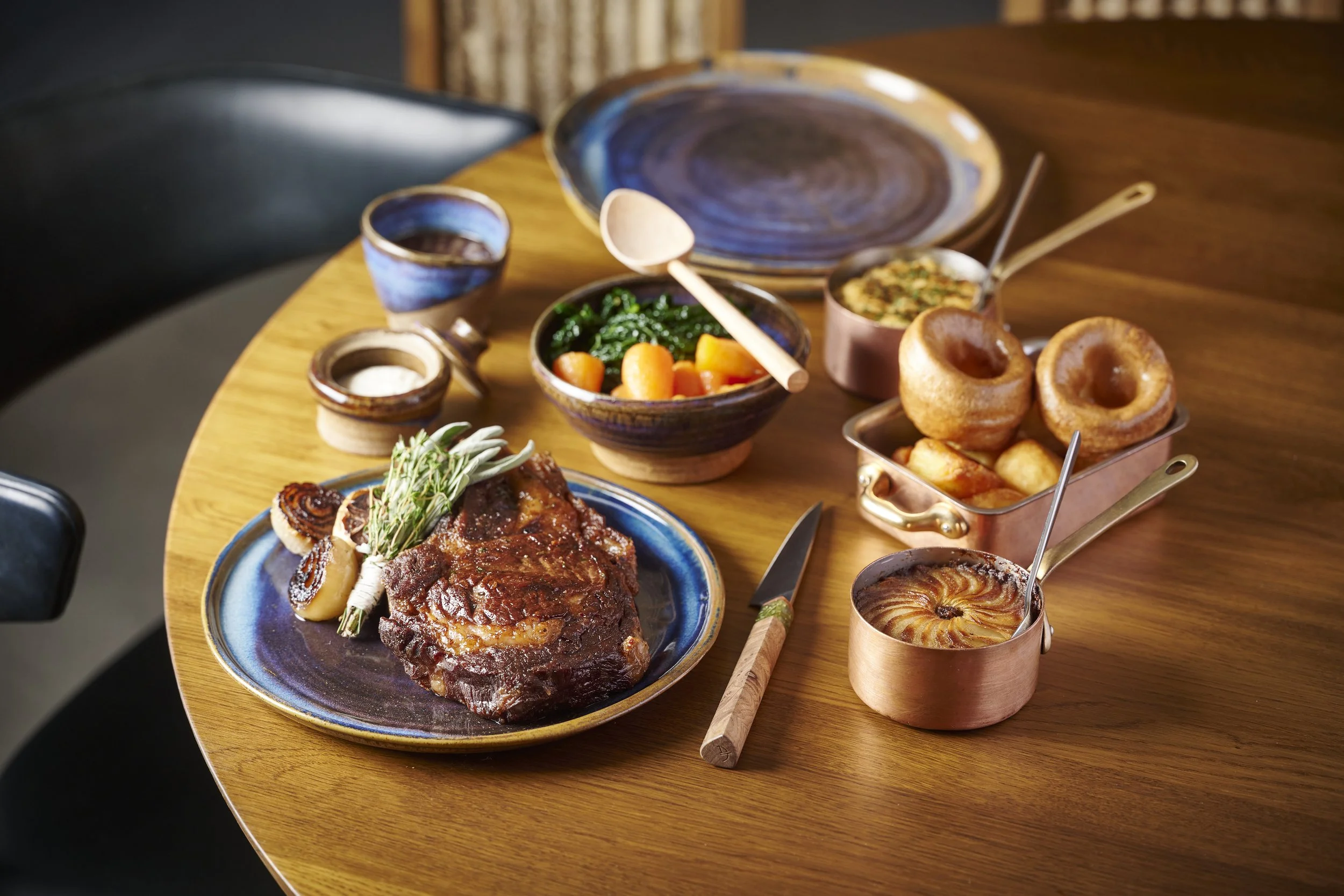 A wooden table with various dishes including a large piece of cooked meat with rosemary, a bowl of cooked greens and carrots, baked onion rings, bread pudding, and small bowls of sauces and condiments.