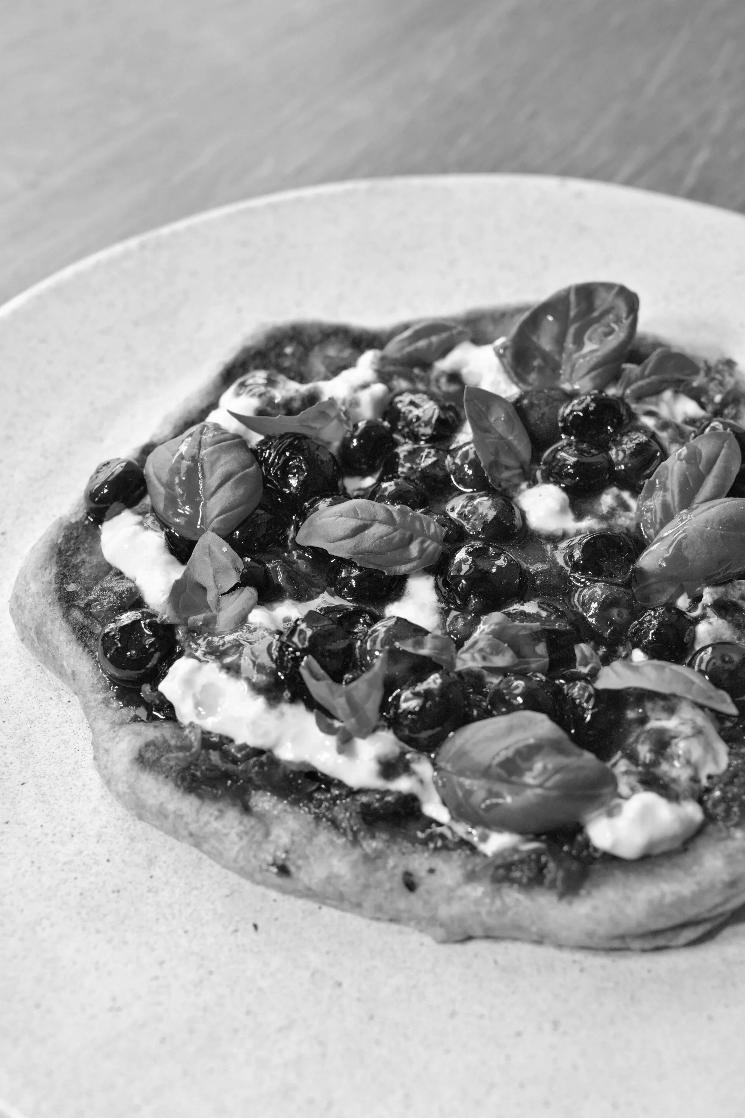 Black and white photo of a pizza with a crust, topped with basil leaves and blueberries, on a white plate.