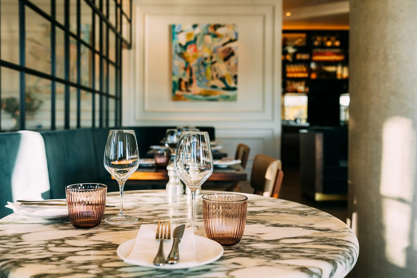 A table set for dining with two empty wine glasses, two pink drinking glasses, a plate with silverware on a napkin, and a salt shaker, in a restaurant with modern decor and artwork on the wall.