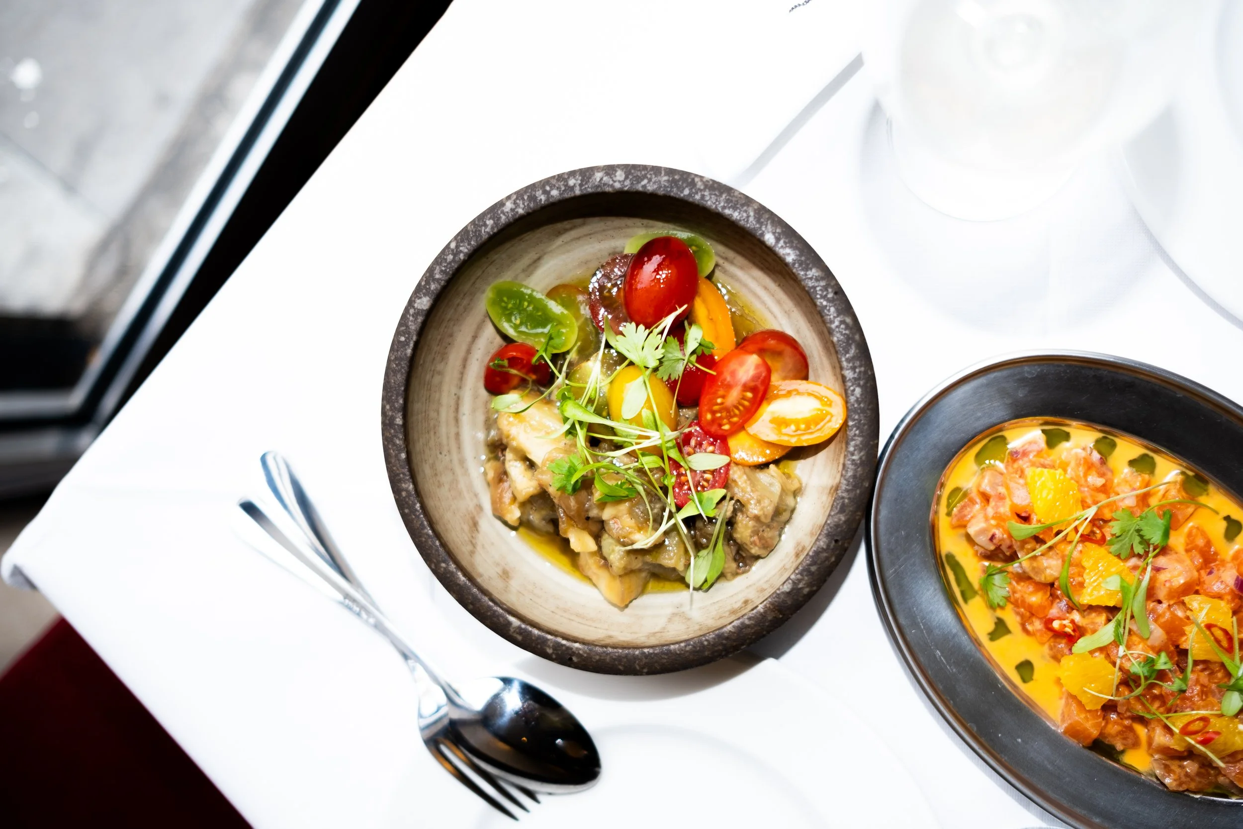 Top view of a table with two dishes: a bowl of chopped vegetables and herbs topped with cherry tomatoes and microgreens, and a dish with creamy seafood and vegetable stew garnished with herbs.