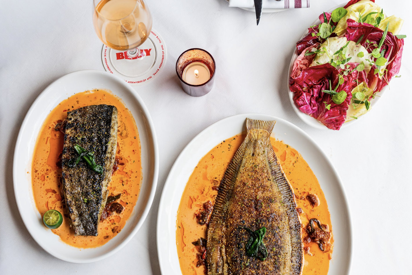 Two plates of grilled fish in orange sauce, garnished with green onions and lime, on a white table with a glass of rosé wine, a lit candle, and a side salad of mixed greens and radicchio.