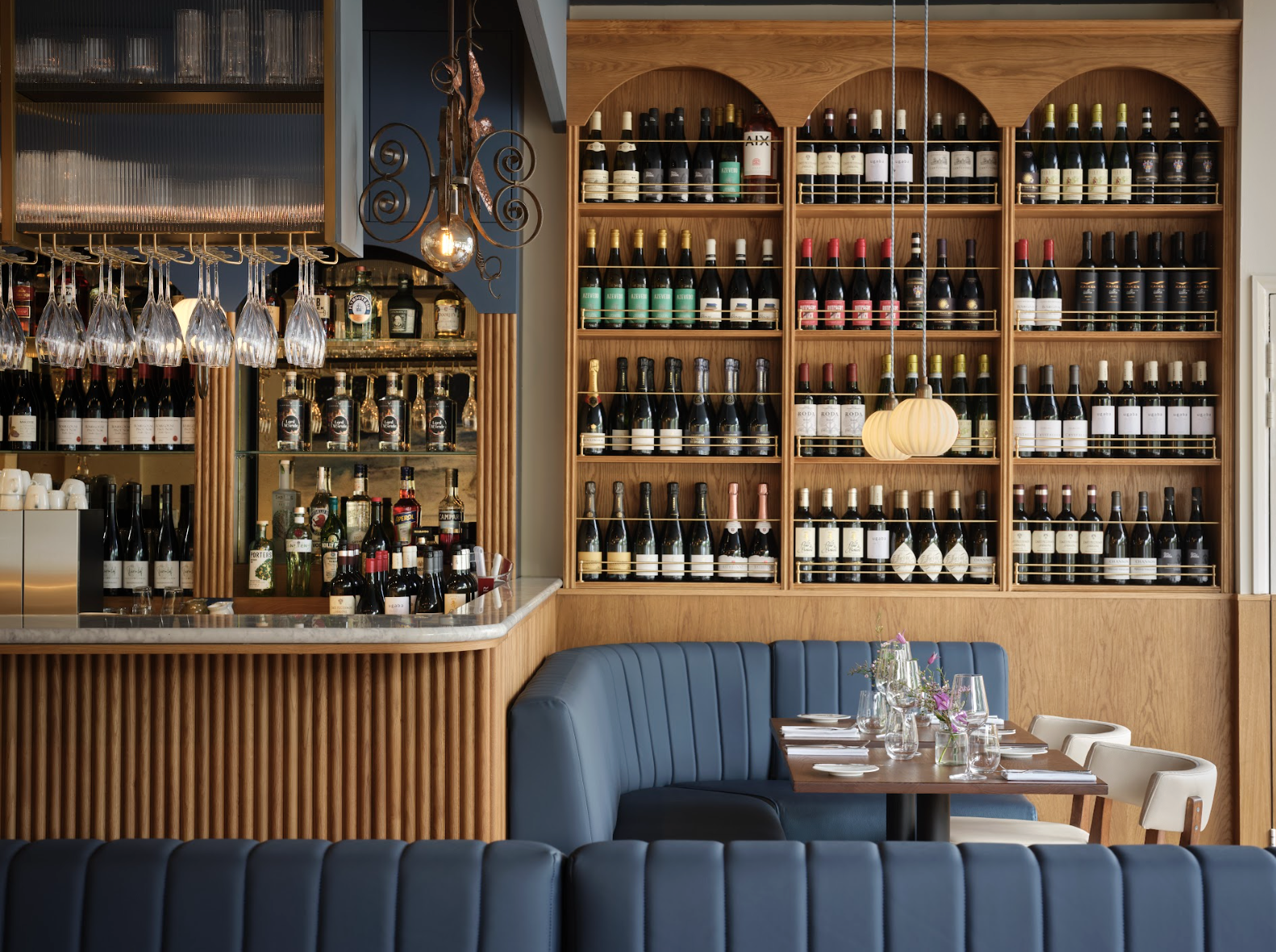 Interior of a restaurant or bar with a wooden wine display, a marble-topped bar counter, hanging glassware, and a blue upholstered booth next to a table set with glassware and a small flower arrangement.
