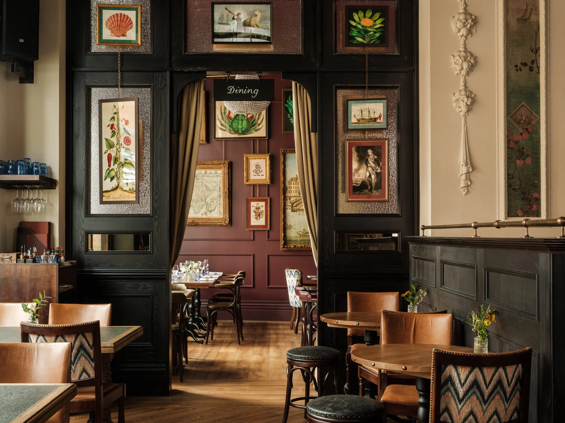Interior of a restaurant with framed art on purple and black walls, round tables with chairs, plants, and a sign that says 'Dining' hanging from the ceiling.