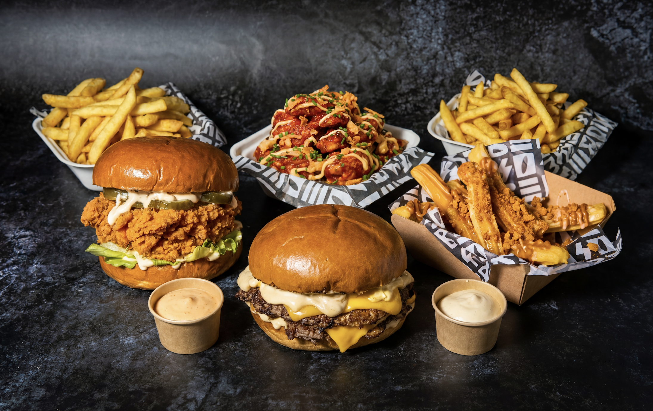 Assorted fast food items including two cheeseburgers with pickles, lettuce, and sauce, two servings of French fries with dipping sauces, a serving of chicken nachos with sauce and chives by The Beefy Boys an Amy Williams Consultancy Client