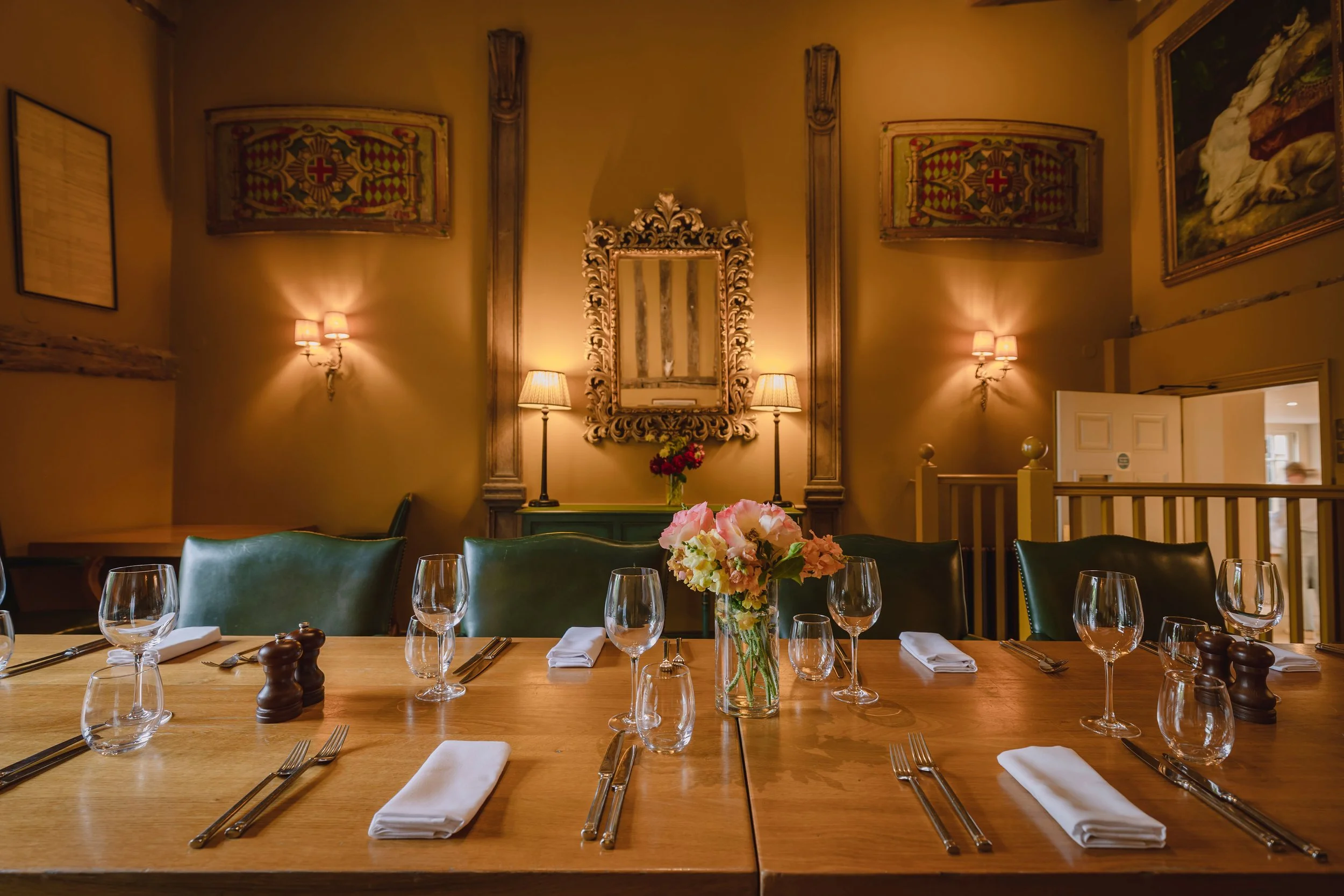 A dining room with a wooden table set with wine glasses, napkins, and cutlery, a floral centerpiece, and a decorated wall with framed art, a mirror, and wall lamps, illuminated with warm lighting.