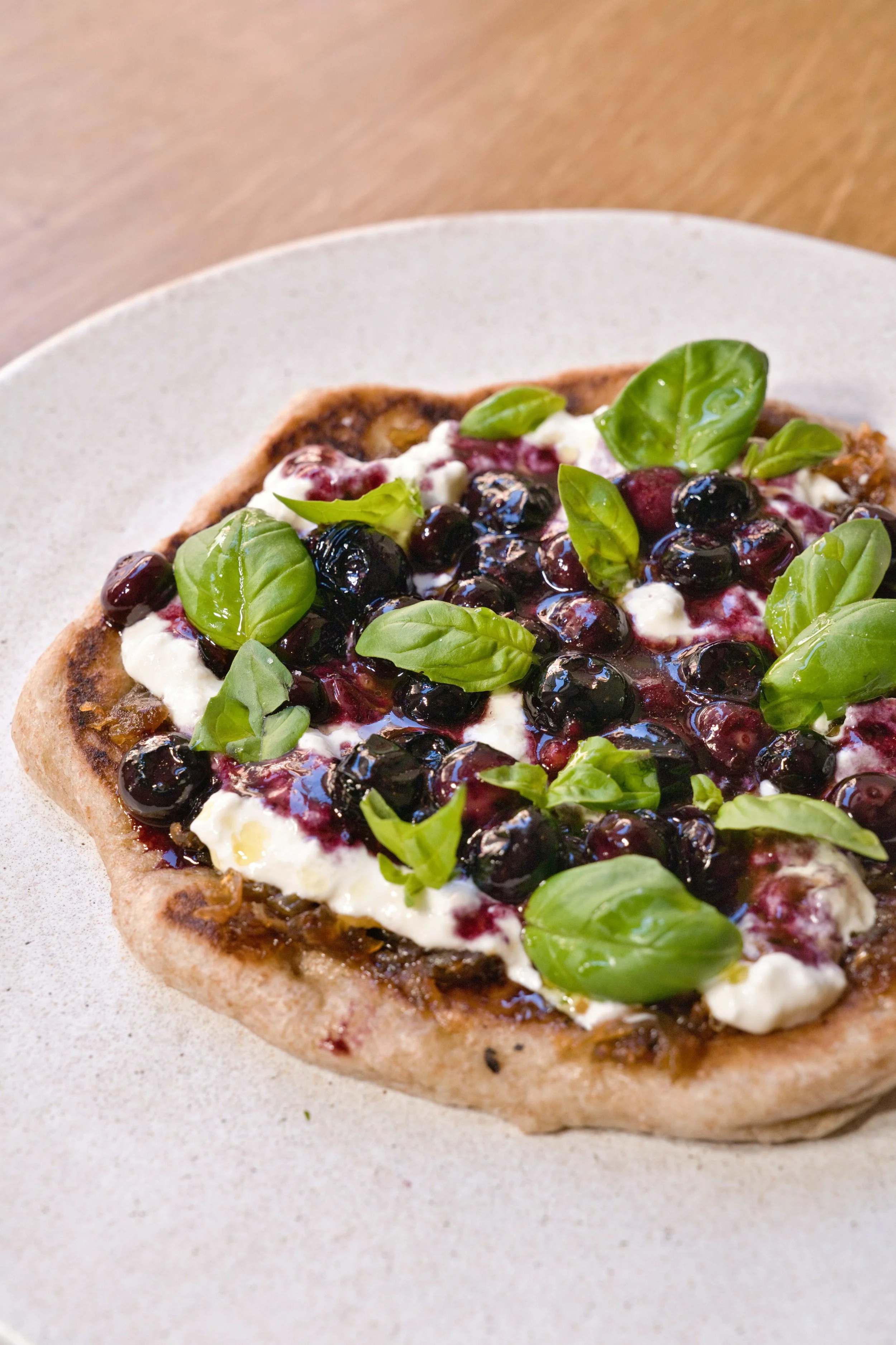 Close-up of a personal-sized pizza topped with blueberries, fresh basil leaves, and dollops of cheese on a white plate.