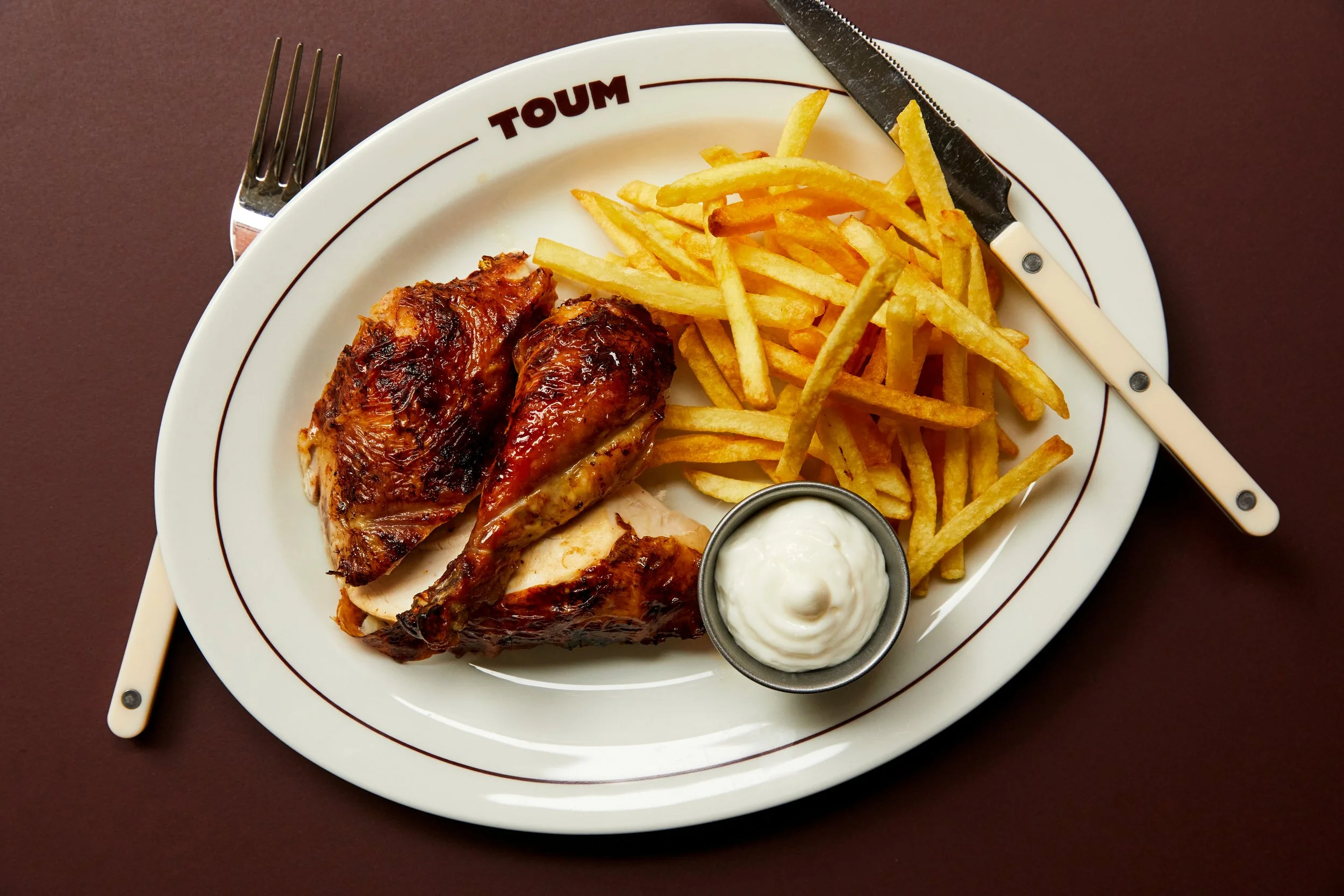 A round plate with the word "TOUM" on the rim, containing grilled chicken, French fries, a small cup of white dipping sauce, a fork, and a knife, on a dark brown table.