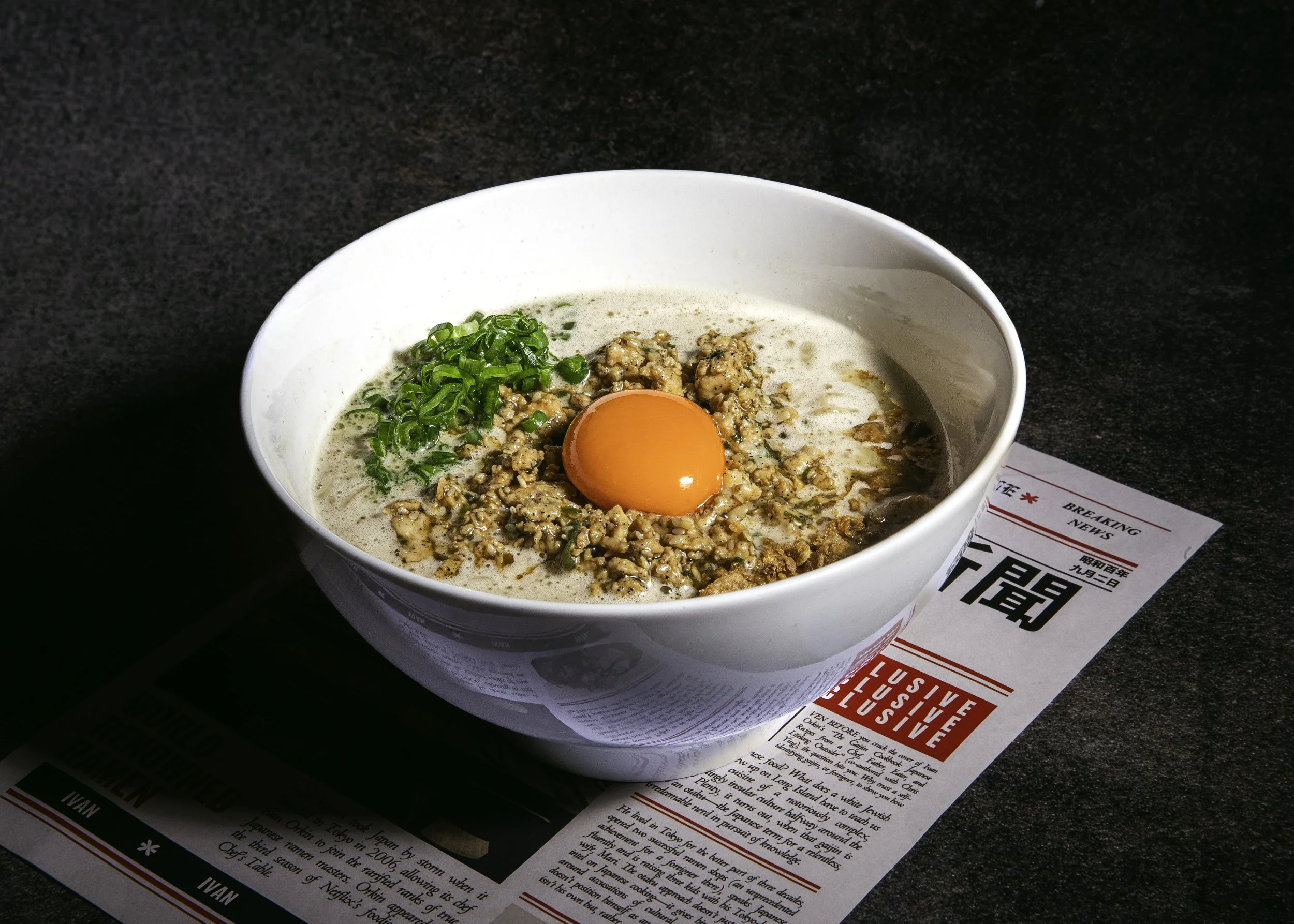 A bowl of ramen with a raw egg yolk, chopped green onions, and seasoned minced meat on top. The bowl rests on a paper food menu on a black tabletop. At Co Thanh Vietnamese an AWC Consultancy client