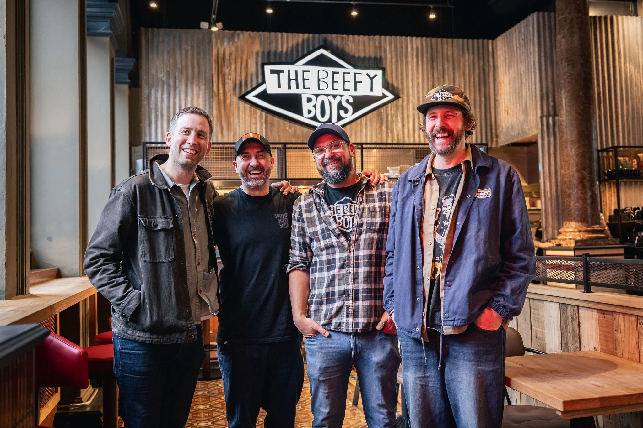 Four men smiling and standing together inside a restaurant, with a sign that reads "The Beefy Boys" behind them. The Beefy Boys are a huge success story and one of Amy Williams Consultancy's biggest growing food brands