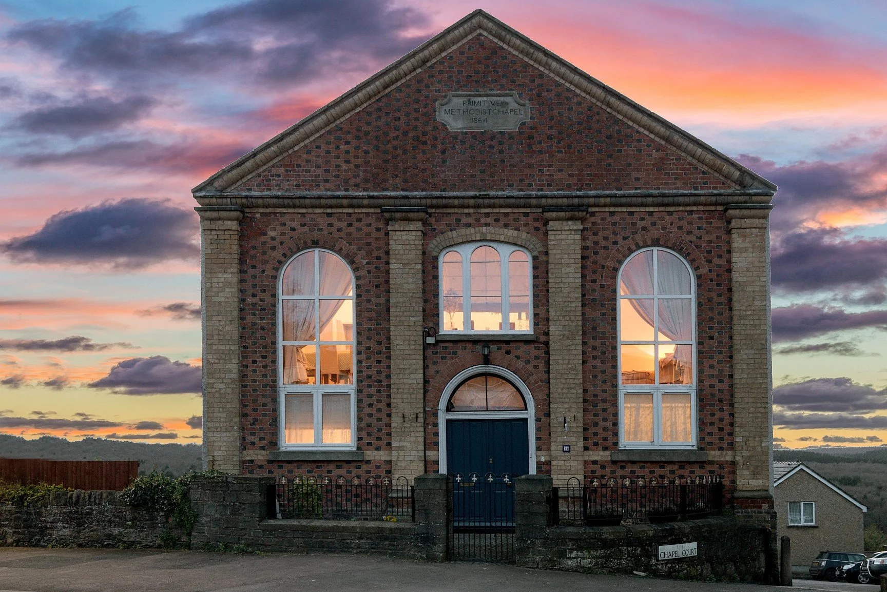 A brick church building with three tall arched windows reflecting a sunset sky with pink and purple clouds, and a blue door. A sign on the building reads 'Primitive Methodist Chapel 1864', and there is a small fence in front.