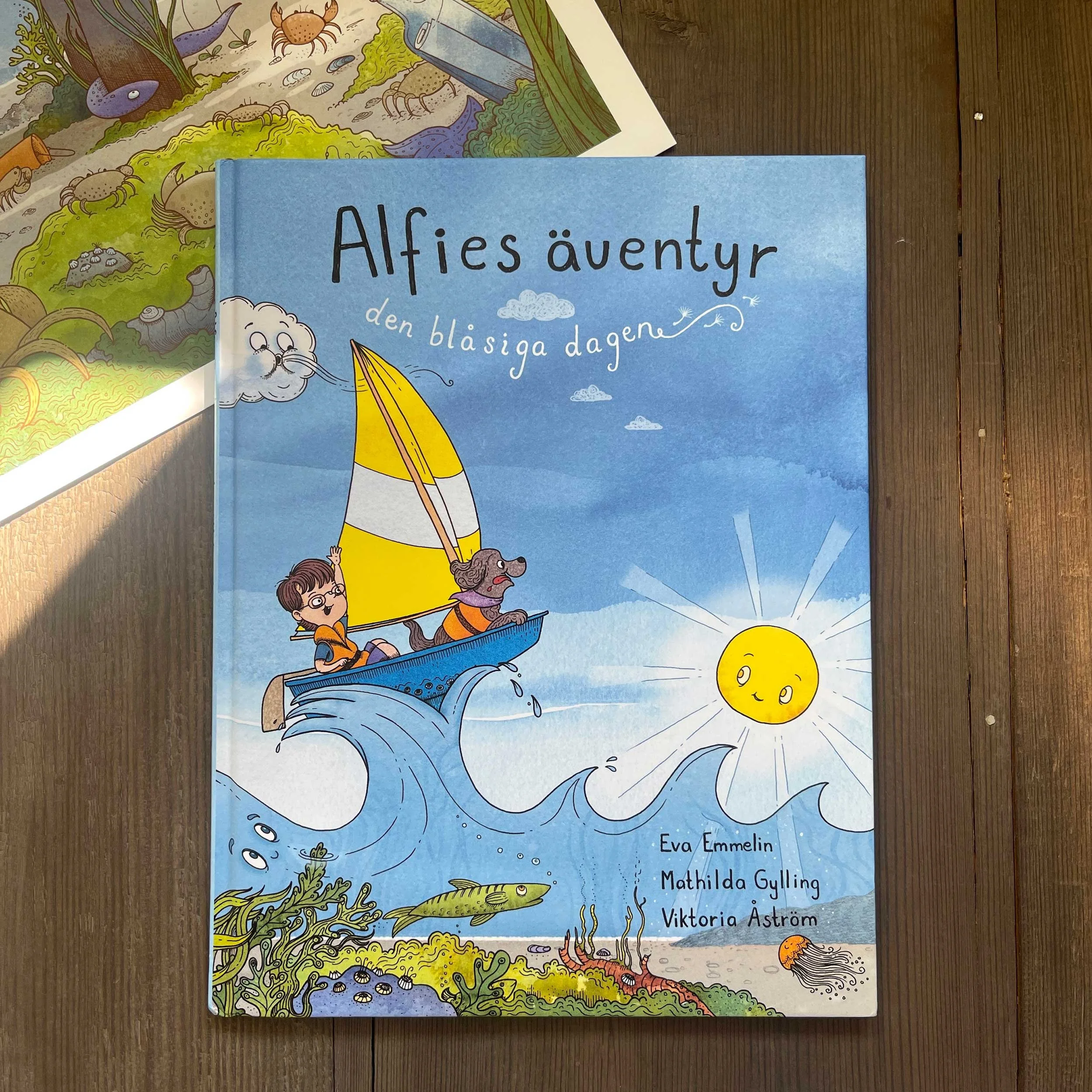 Eolus Vind - Children's book - a boy and a dog in a boat sailing on a wave, with a smiling sun in the sky and underwater plants, fish, and sea creatures below. Illustrations by Viktoria Åström