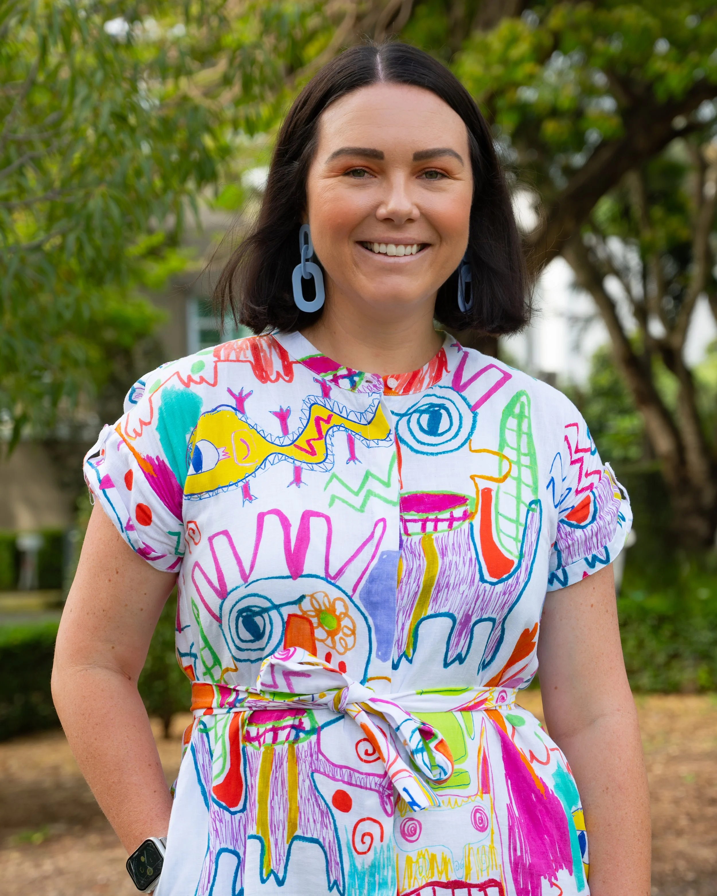 A woman smiling outdoors, wearing a colorful, abstract patterned dress and blue earrings, with trees in the background.
