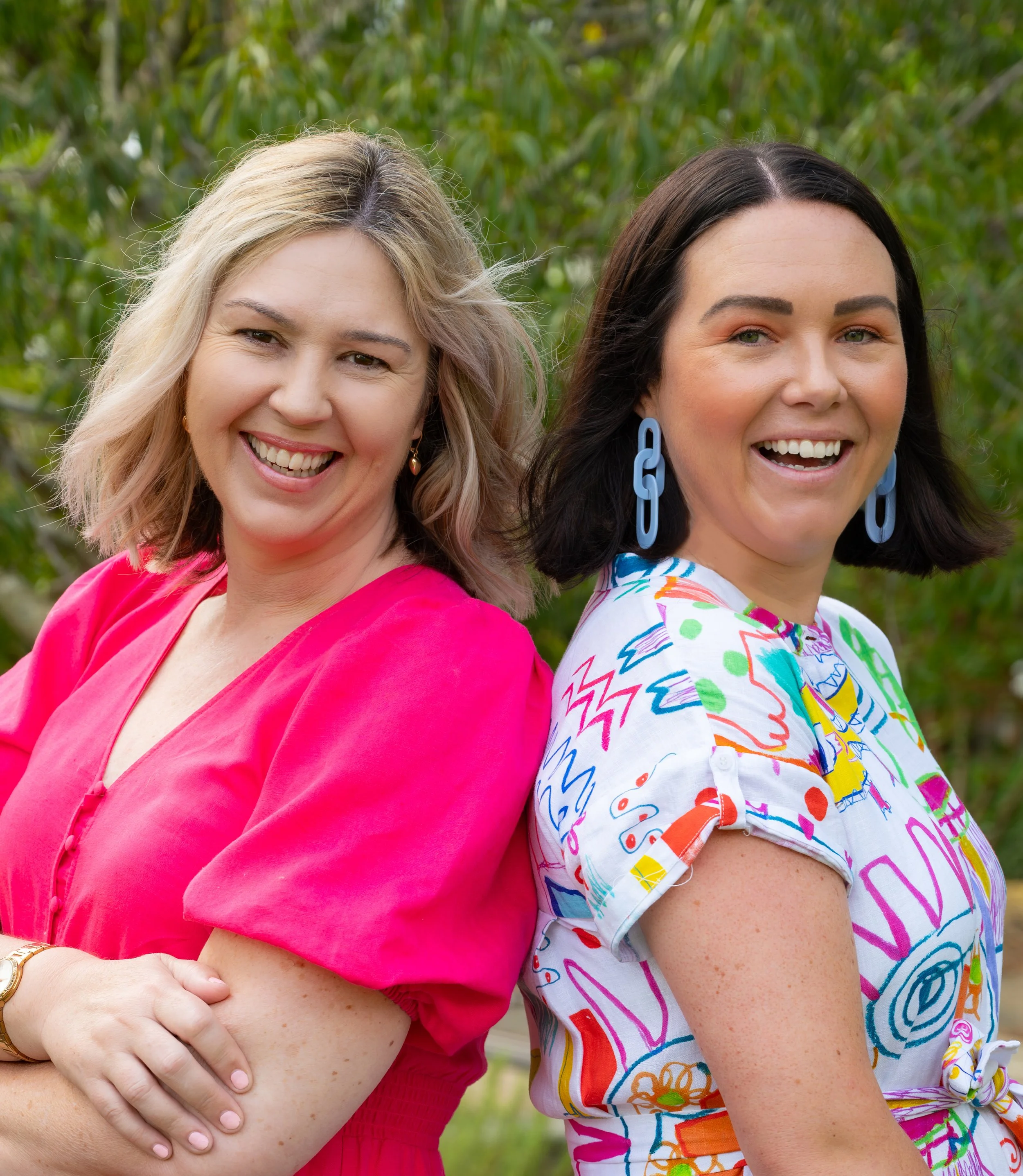 Two smiling women standing back to back outdoors, one wearing a pink blouse and the other wearing a colorful patterned top and blue earrings, with green foliage in the background.