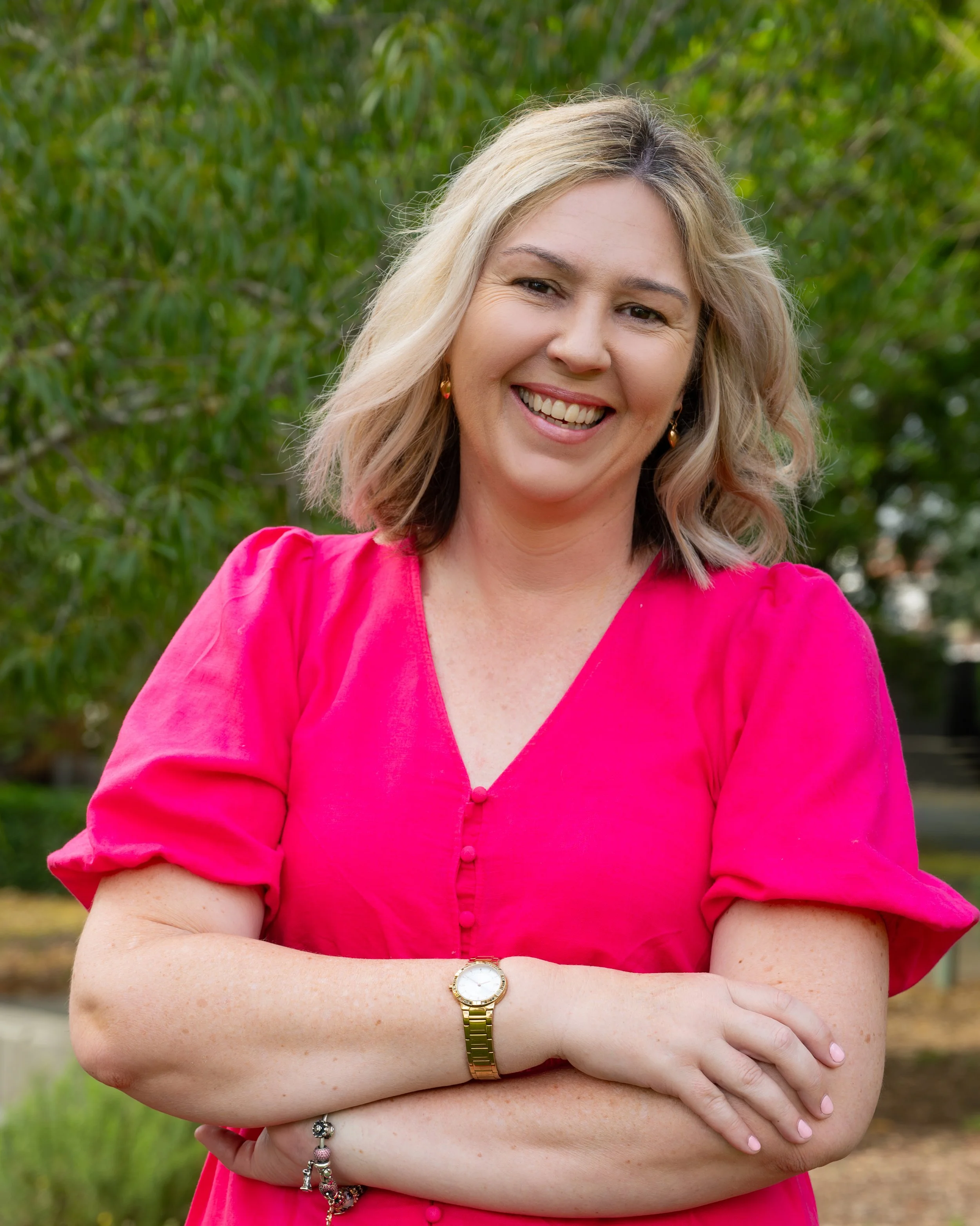 A woman with blonde hair smiling in a pink dress outdoors, with green trees in the background.