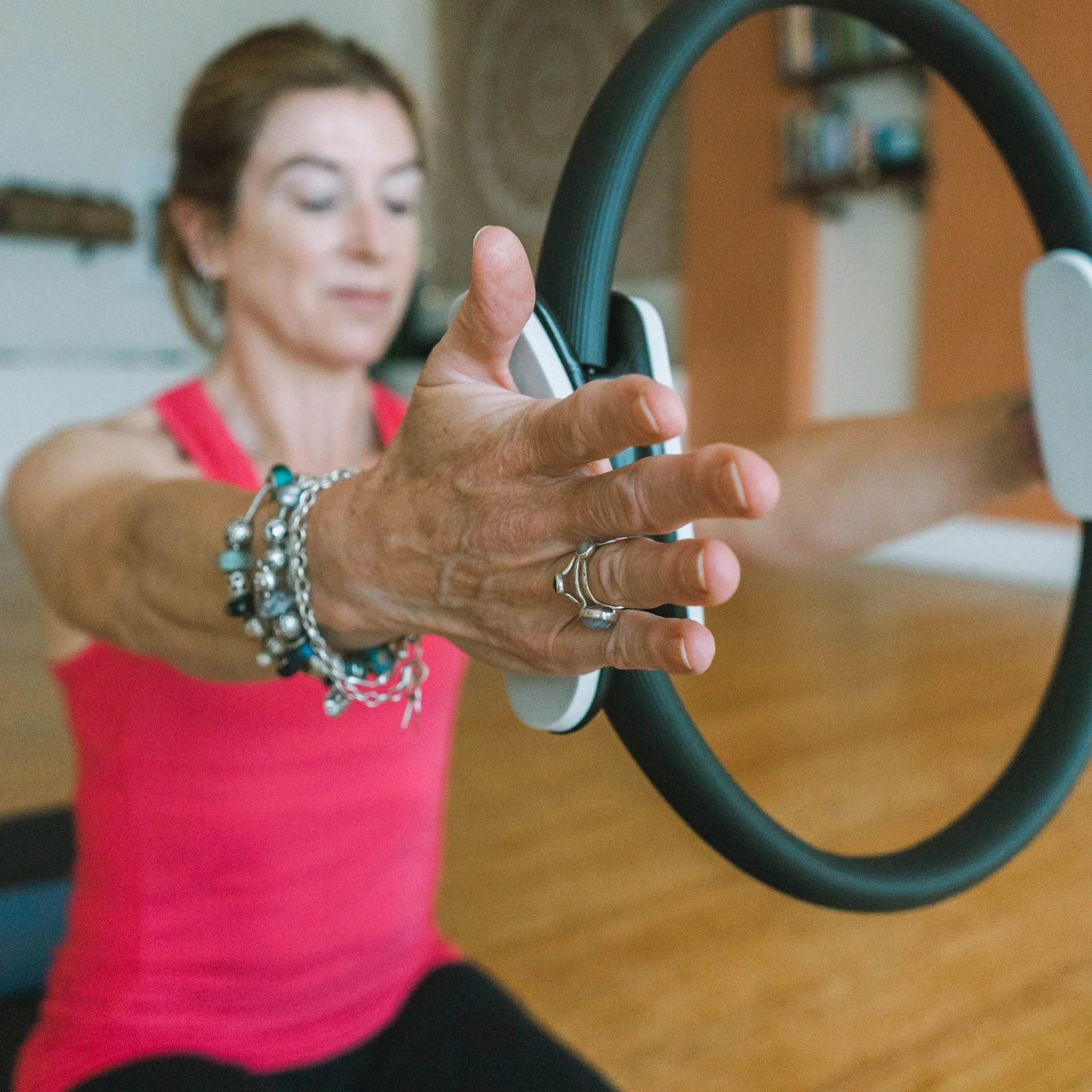 A woman in a pink top is sitting on the floor, reaching out with both hands while holding a Pilates Ring.