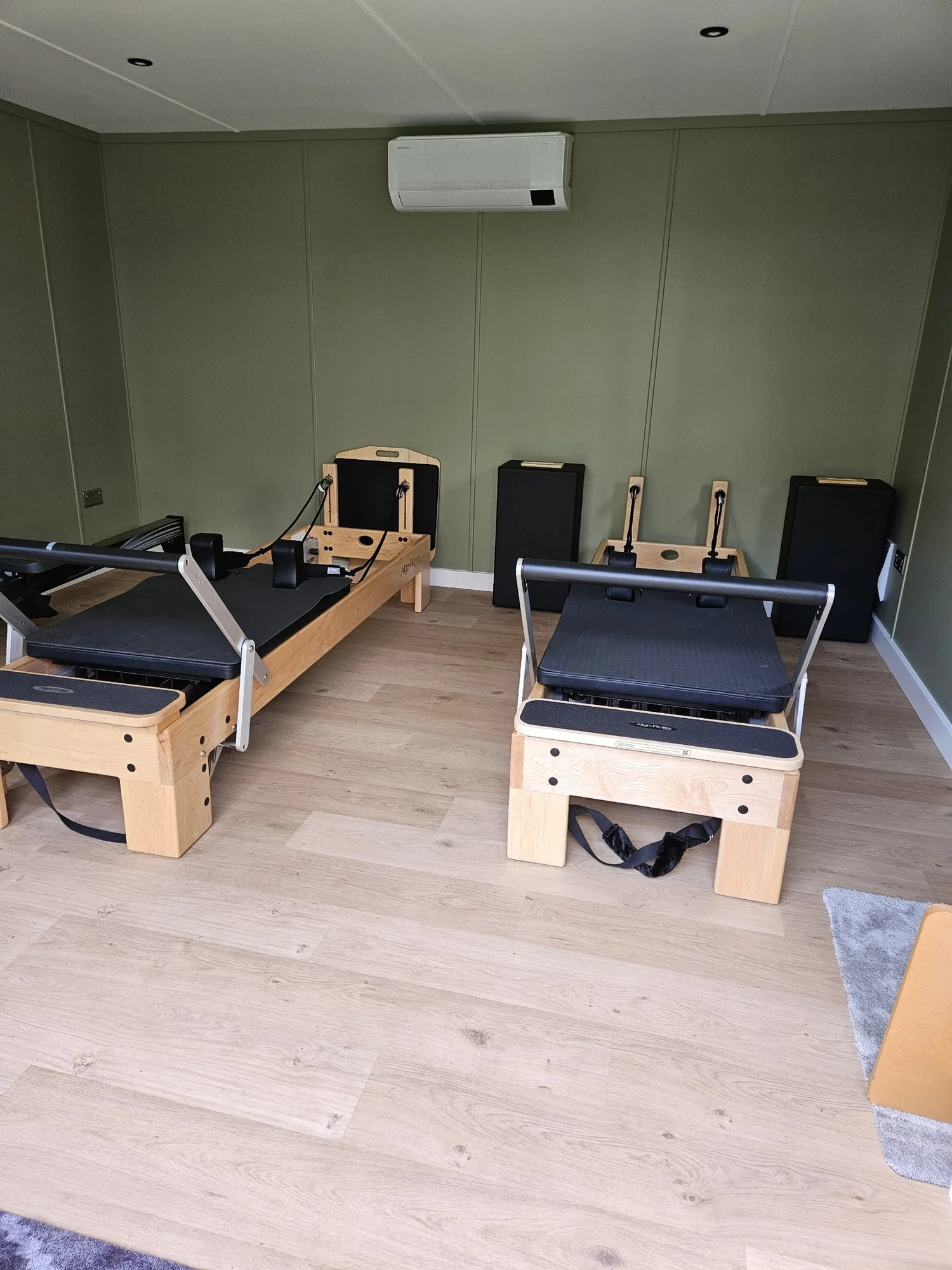 Two wooden Pilates reformer machines with black padding, positioned in a room with light wood flooring and green walls, with a wall-mounted air conditioning unit above.