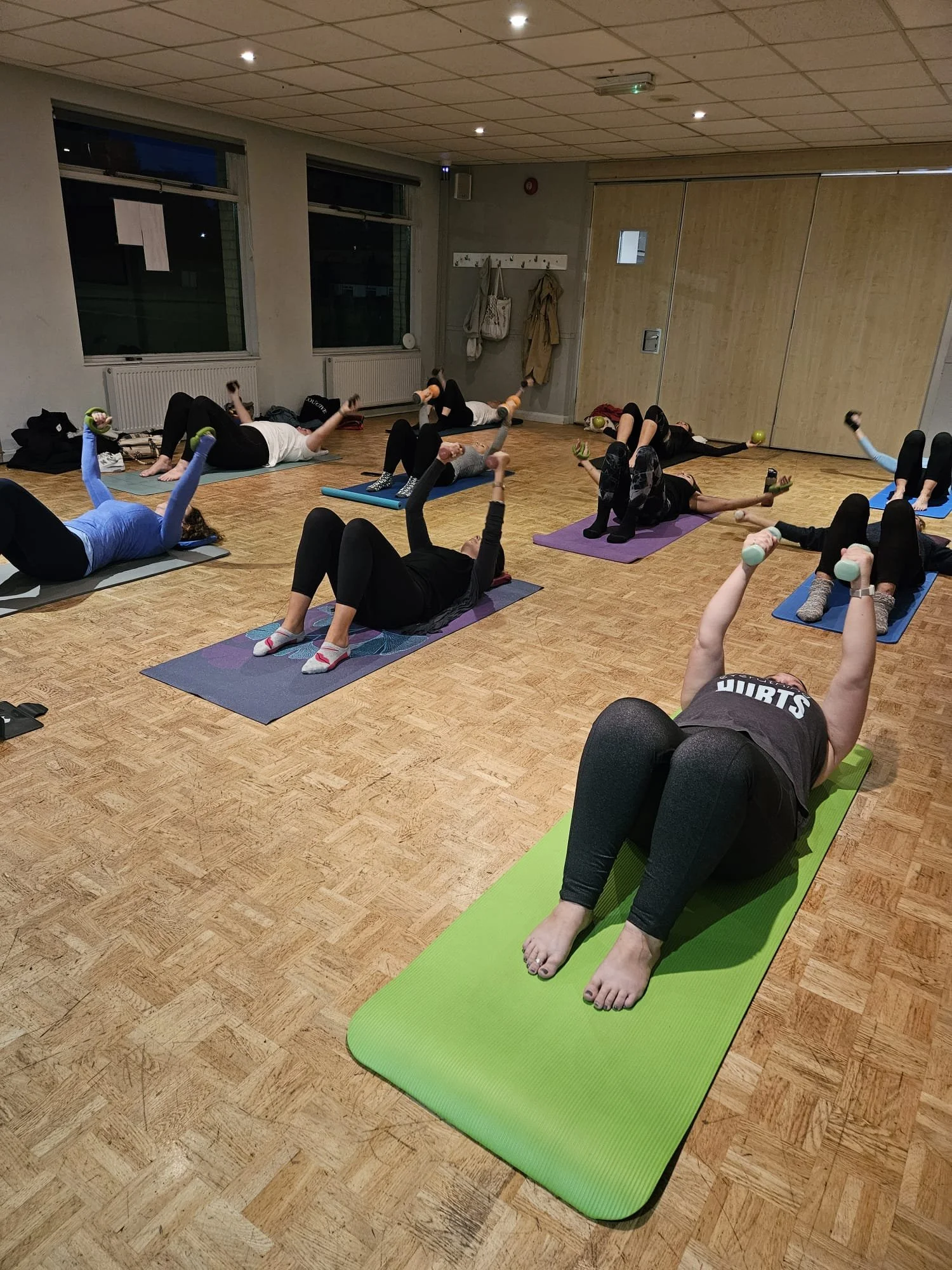 Group exercise class in a studio with people lying on mats, lifting small weights in the air, practicing strength training.