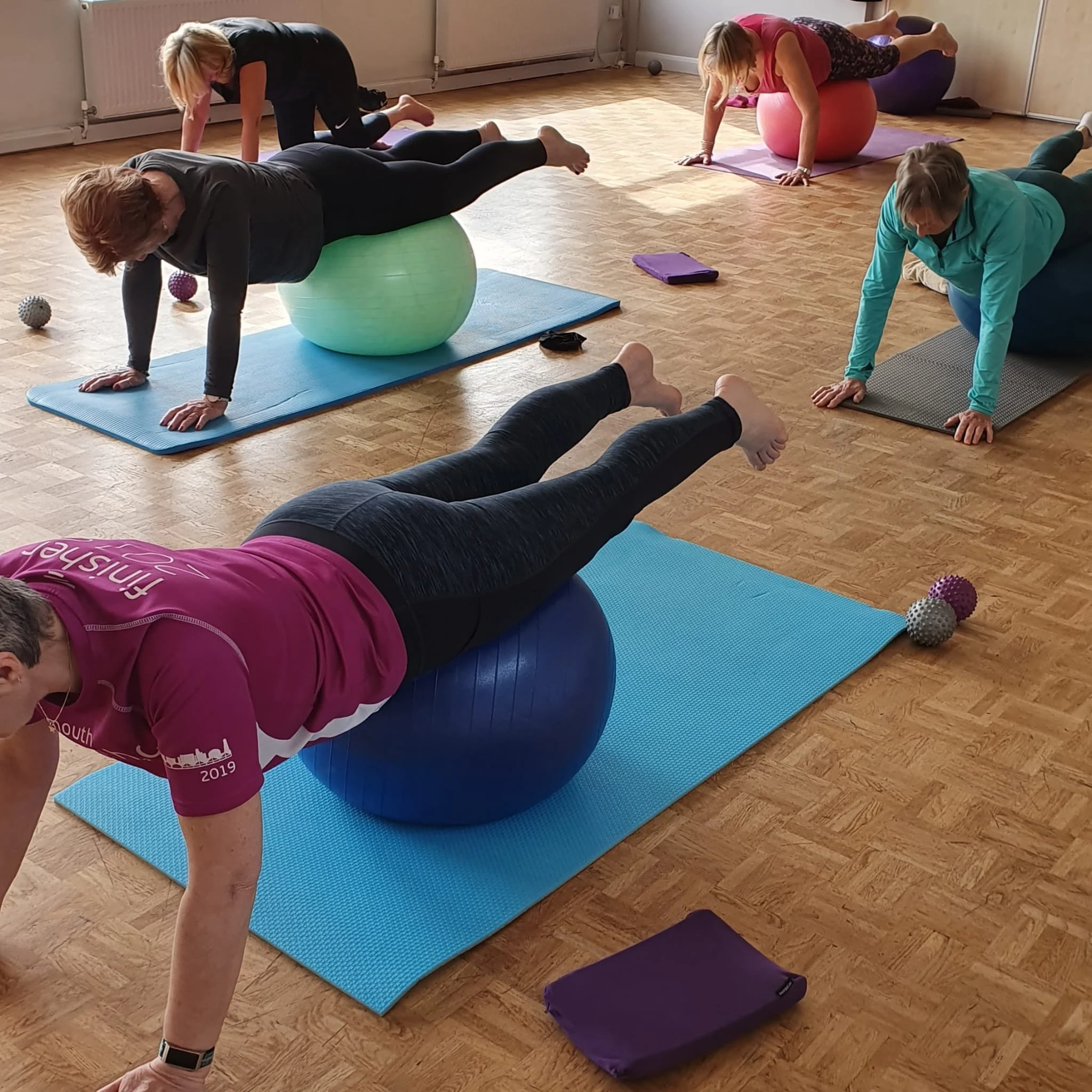 A group of women participating in a fitness class using exercise balls on yoga mats in a studio with wooden floors.