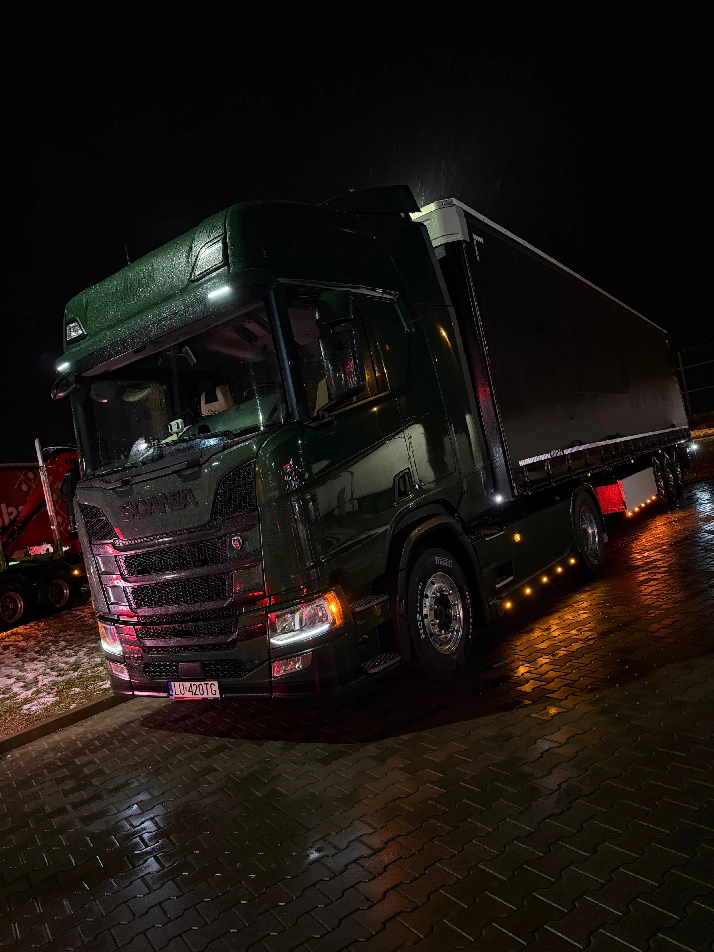 Nighttime photo of a dark green semi-truck parked on brick pavement with wet surface, with snow patches visible on the ground, headlights on, and surrounding darkness.