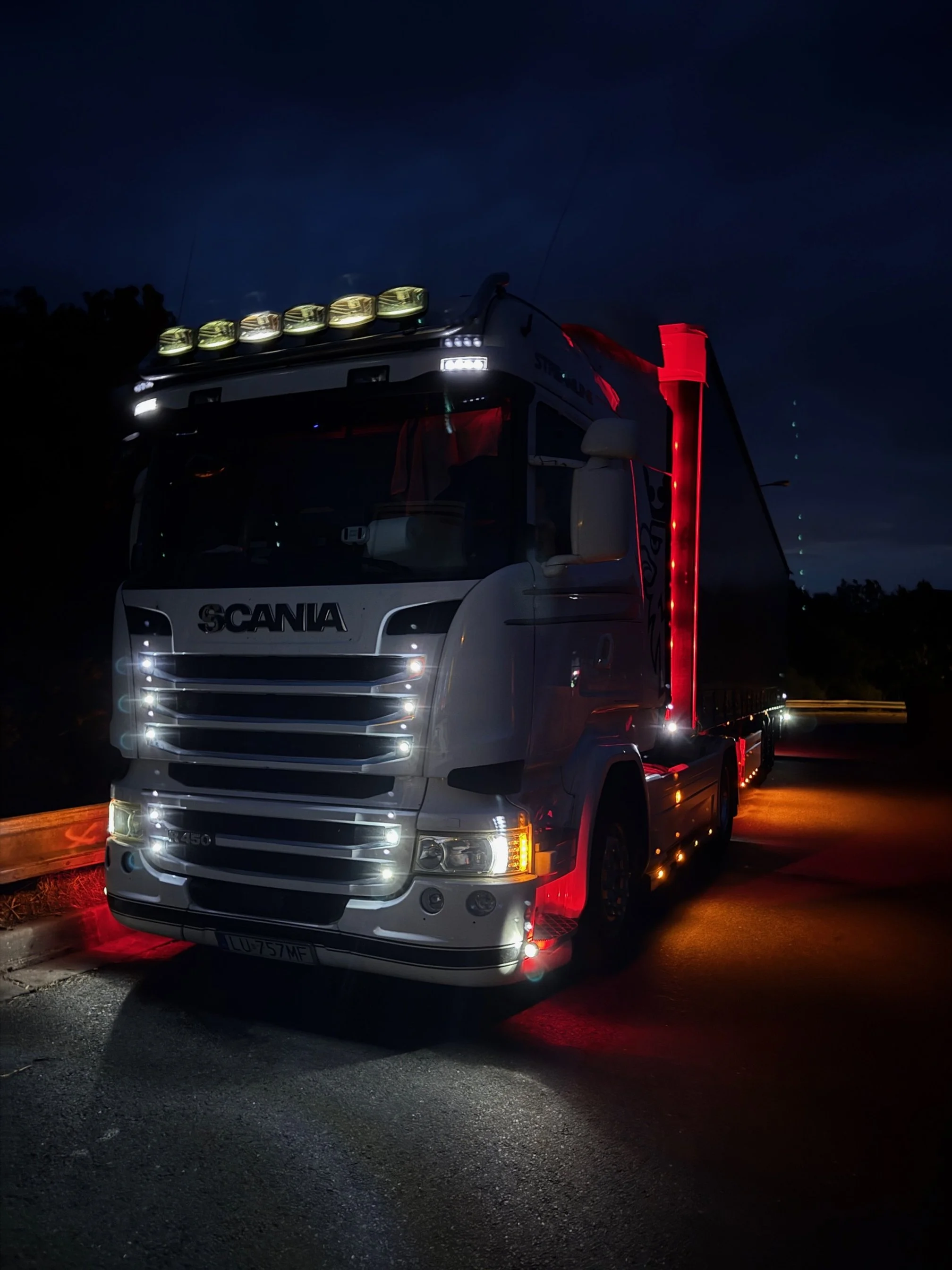 Nighttime photo of a white Scania truck with illuminated lights, parked on the side of the road with a dark sky in the background.