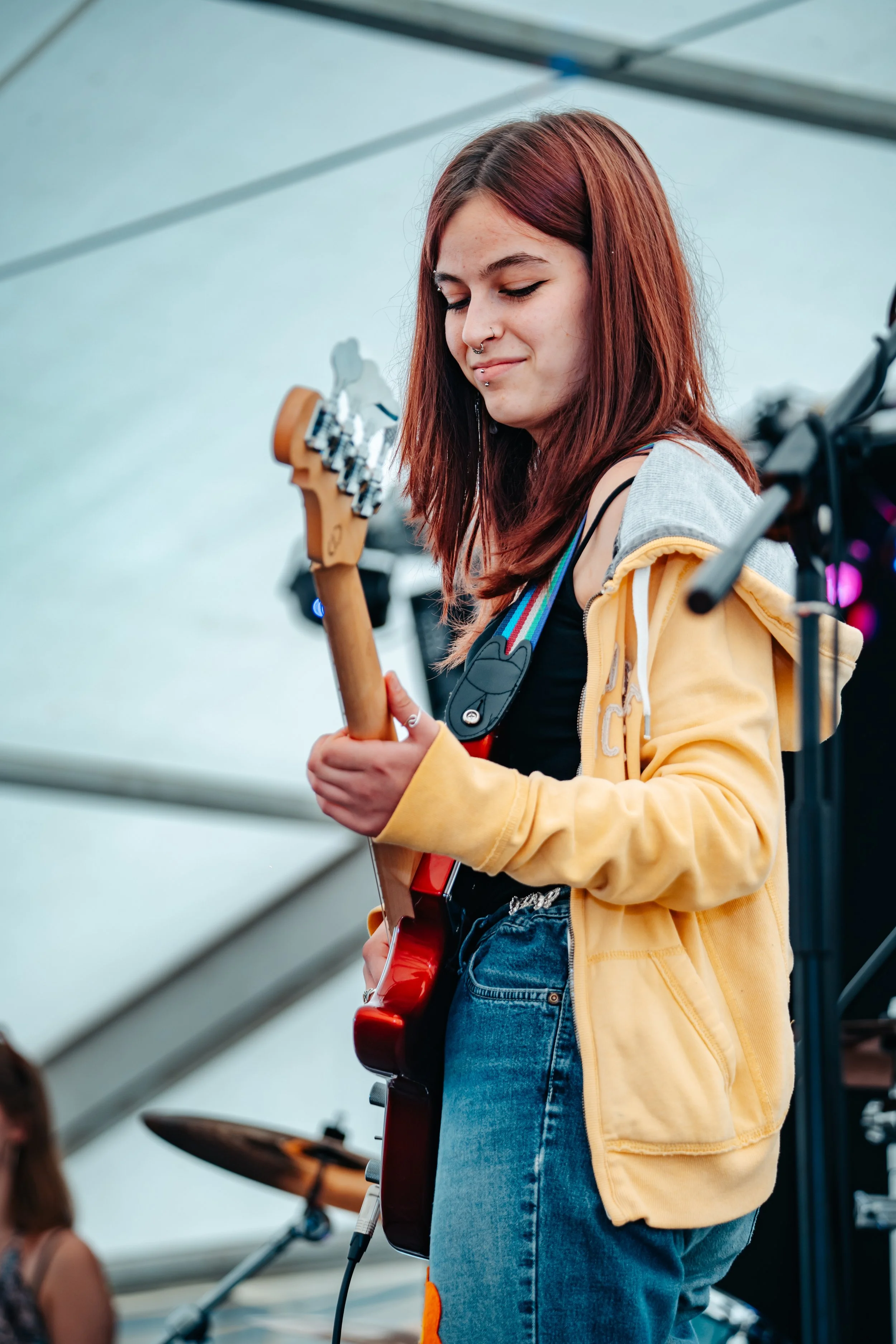 A young woman with red hair playing an electric guitar on stage, wearing a yellow hoodie and jeans, with a focused expression.