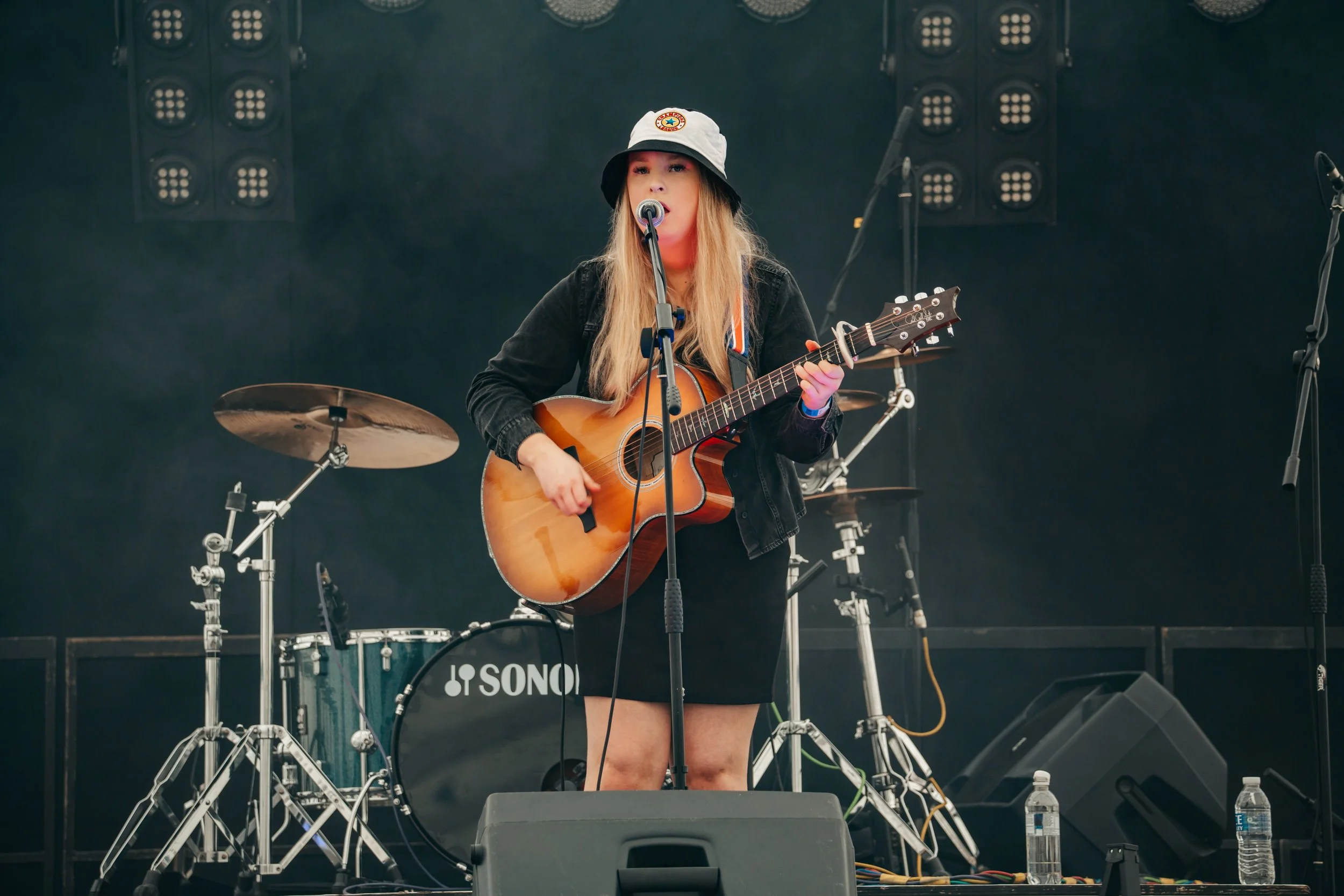 Female musician with long blond hair wearing a black jacket, black skirt, and a bucket hat playing an acoustic guitar on stage with drums and other equipment in the background.