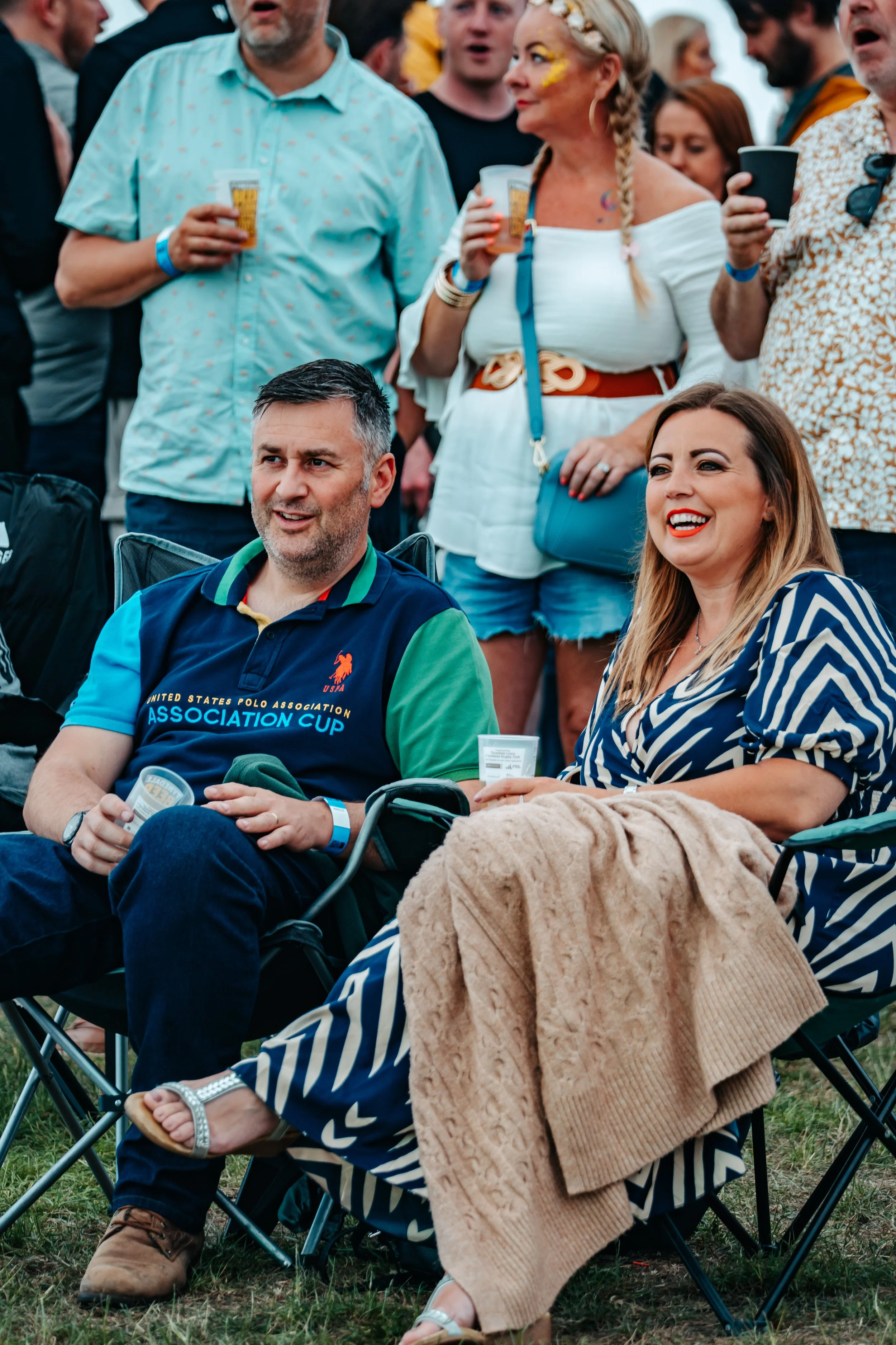 Two people sitting in chairs at an outdoor event, surrounded by standing people; the man wears a colorful polo shirt, and the woman wears a blue and white striped dress, both smiling and holding drinks.