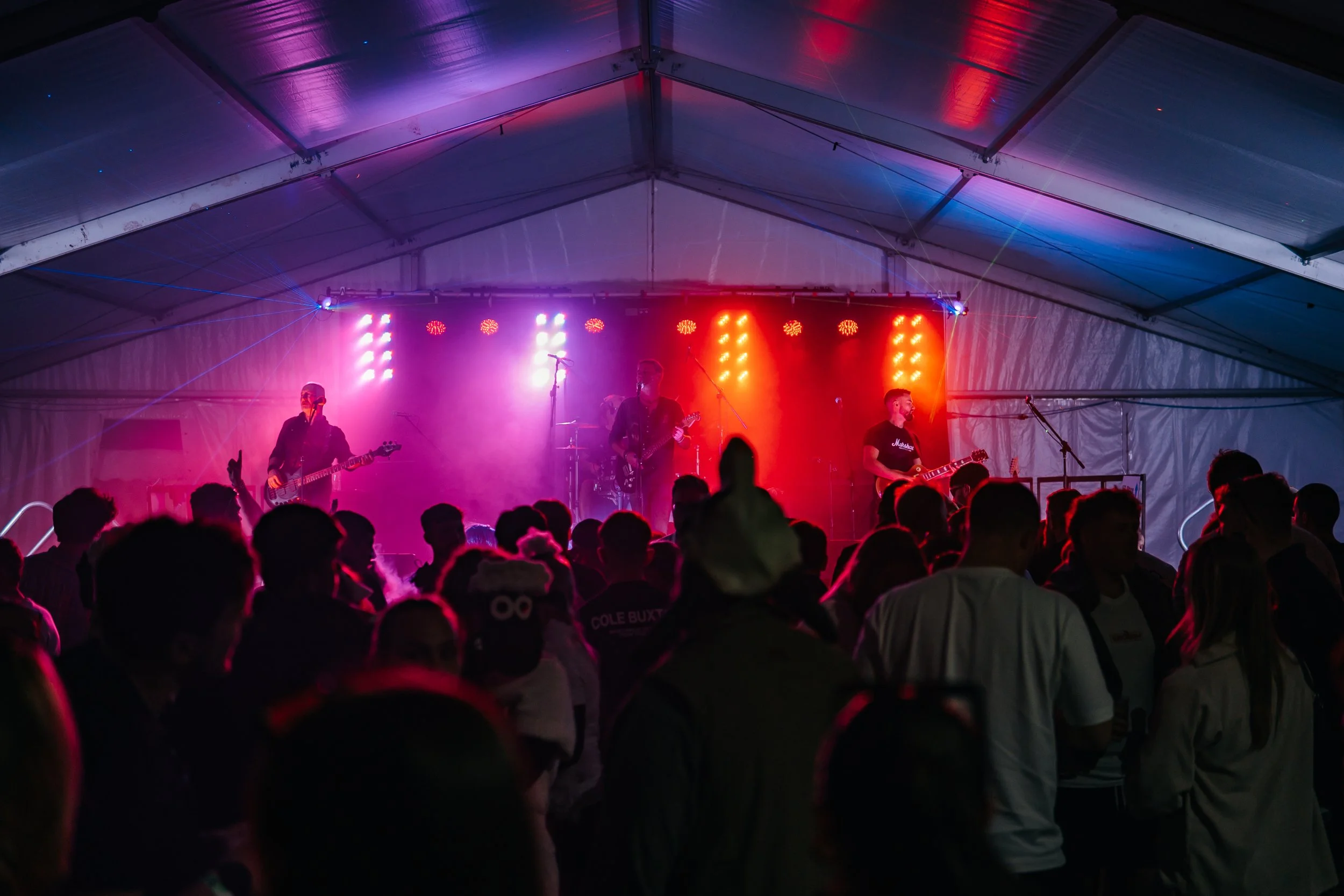 A live concert inside a large tent with a band performing on stage illuminated by colorful lights, and an audience watching.