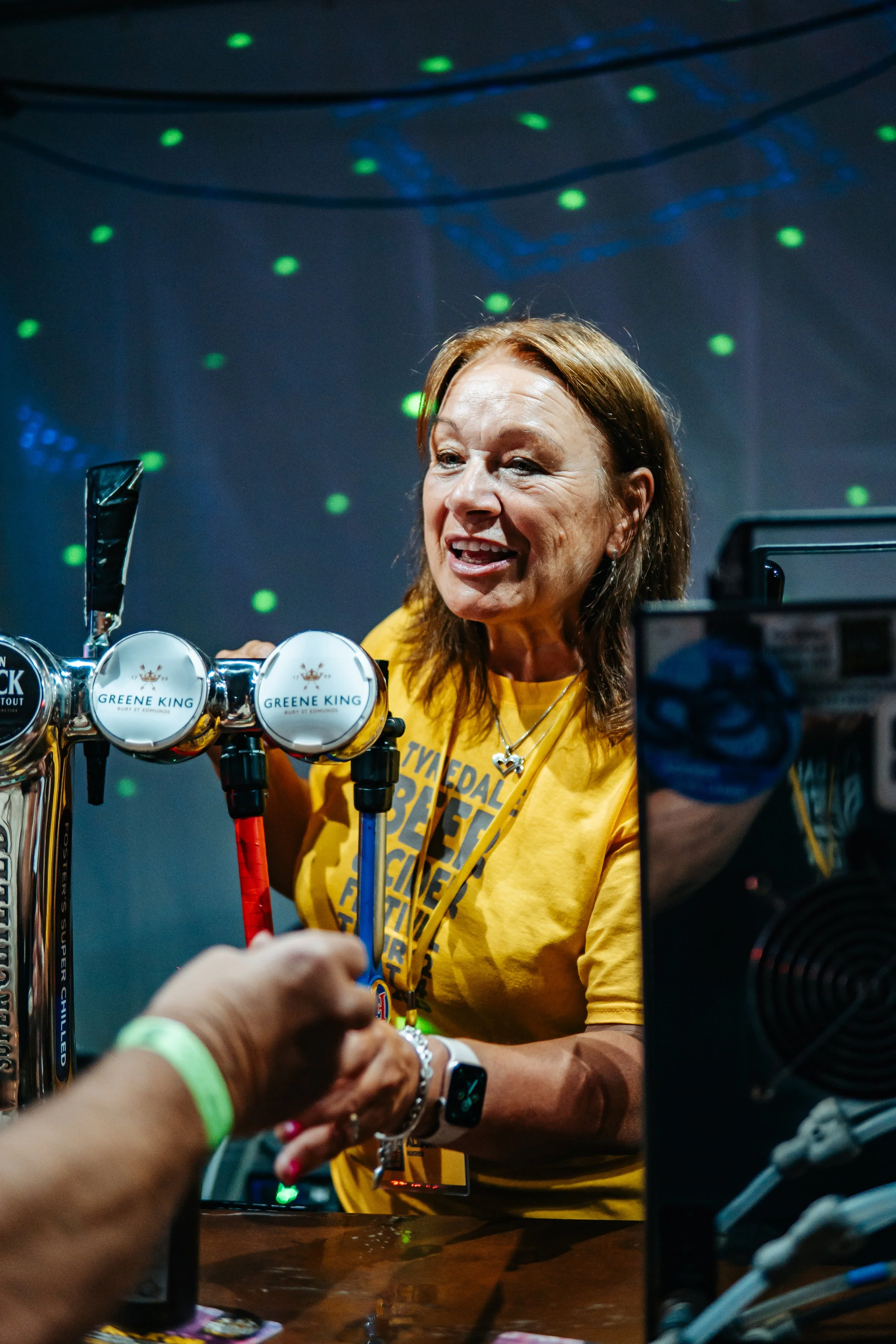 A woman with reddish-brown hair wearing a yellow shirt and a necklace with a heart pendant serving drinks at a bar with a neon-lit backdrop