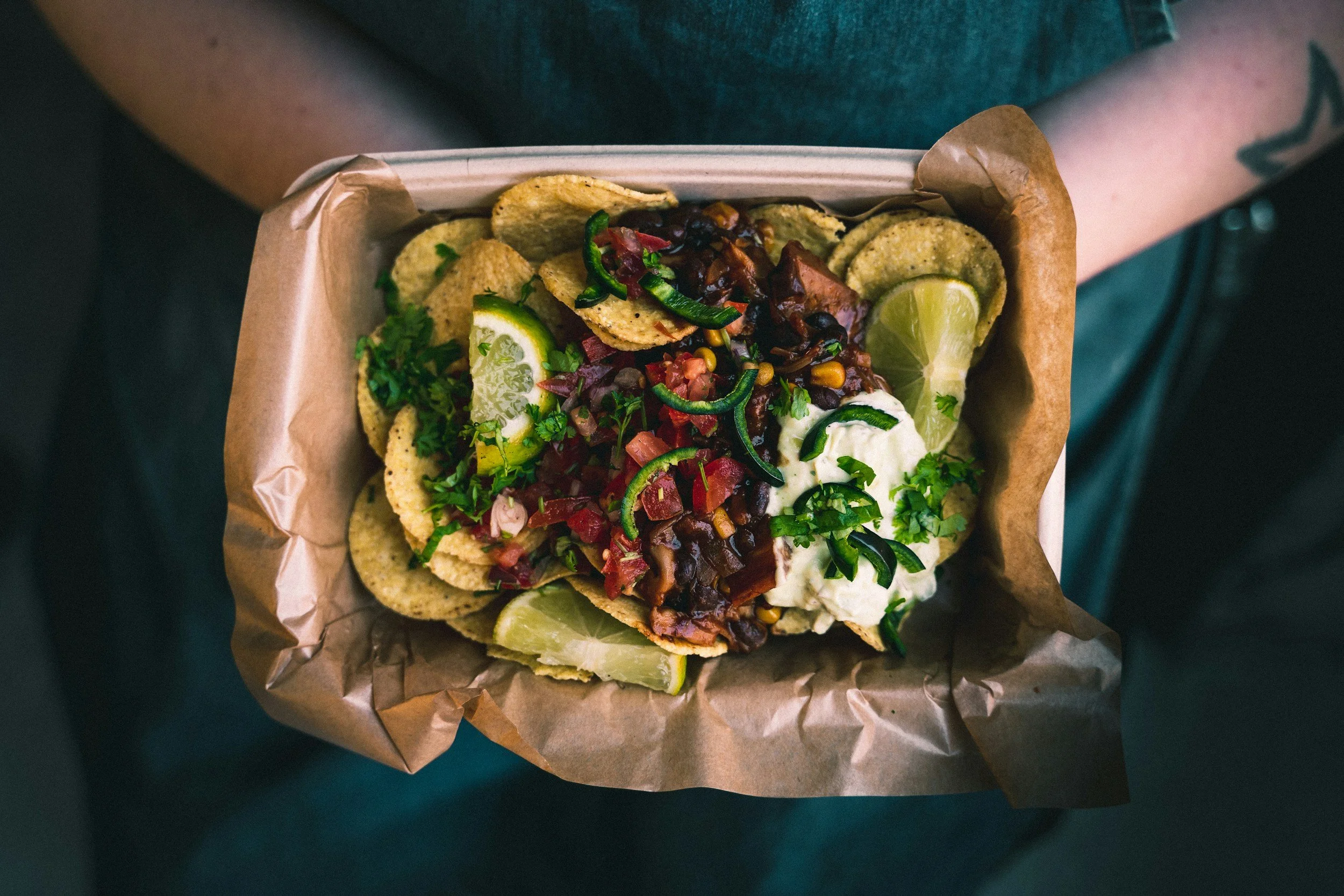 Bowl of nachos topped with black beans, chopped tomatoes, chopped green onions, salsa, shredded cheese, sour cream, chopped cilantro, and sliced lime wedges, with crispy potato chips around the edges.