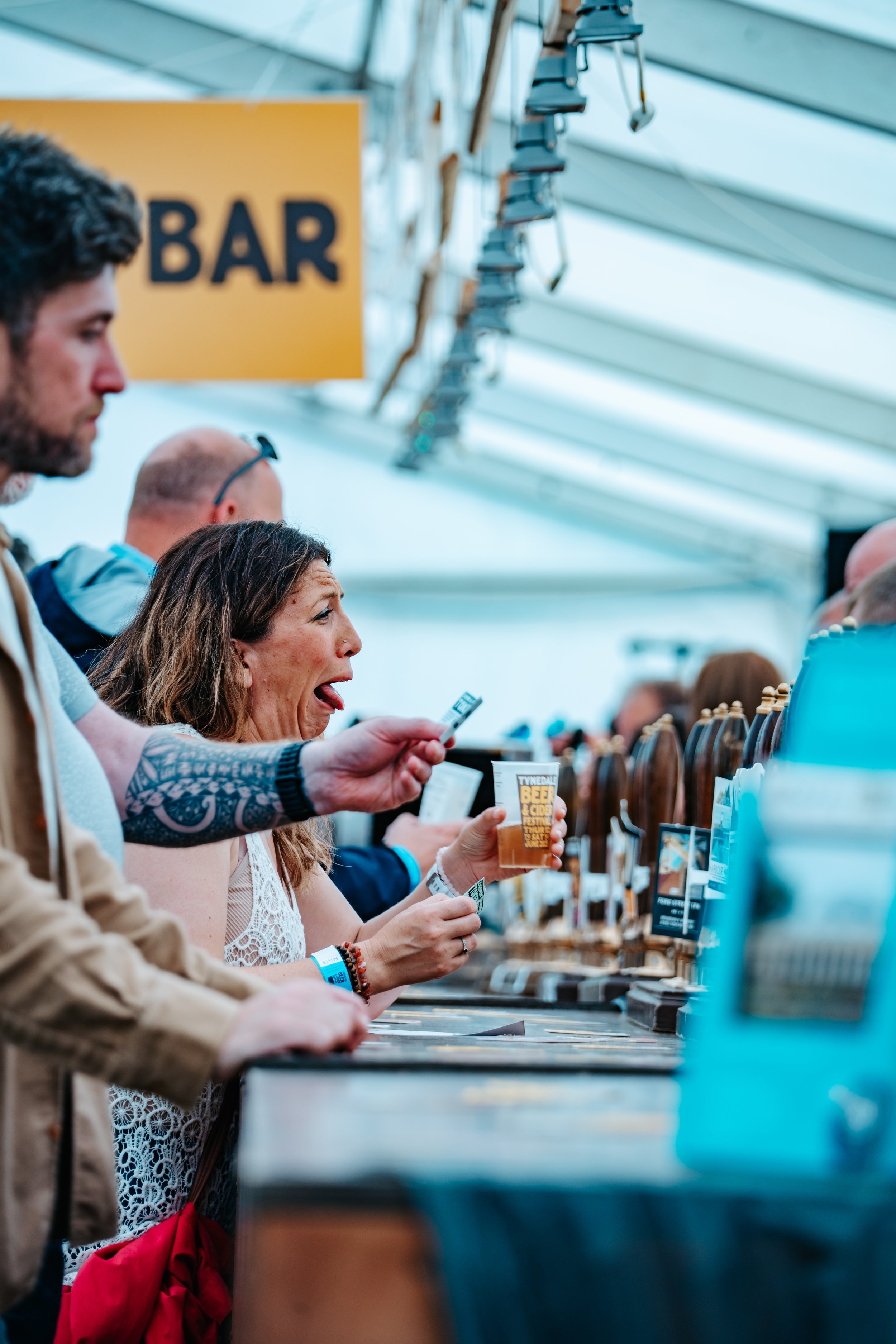 People at a bar counter ordering drinks, one woman holding a beer glass with a sign for a beer festival.