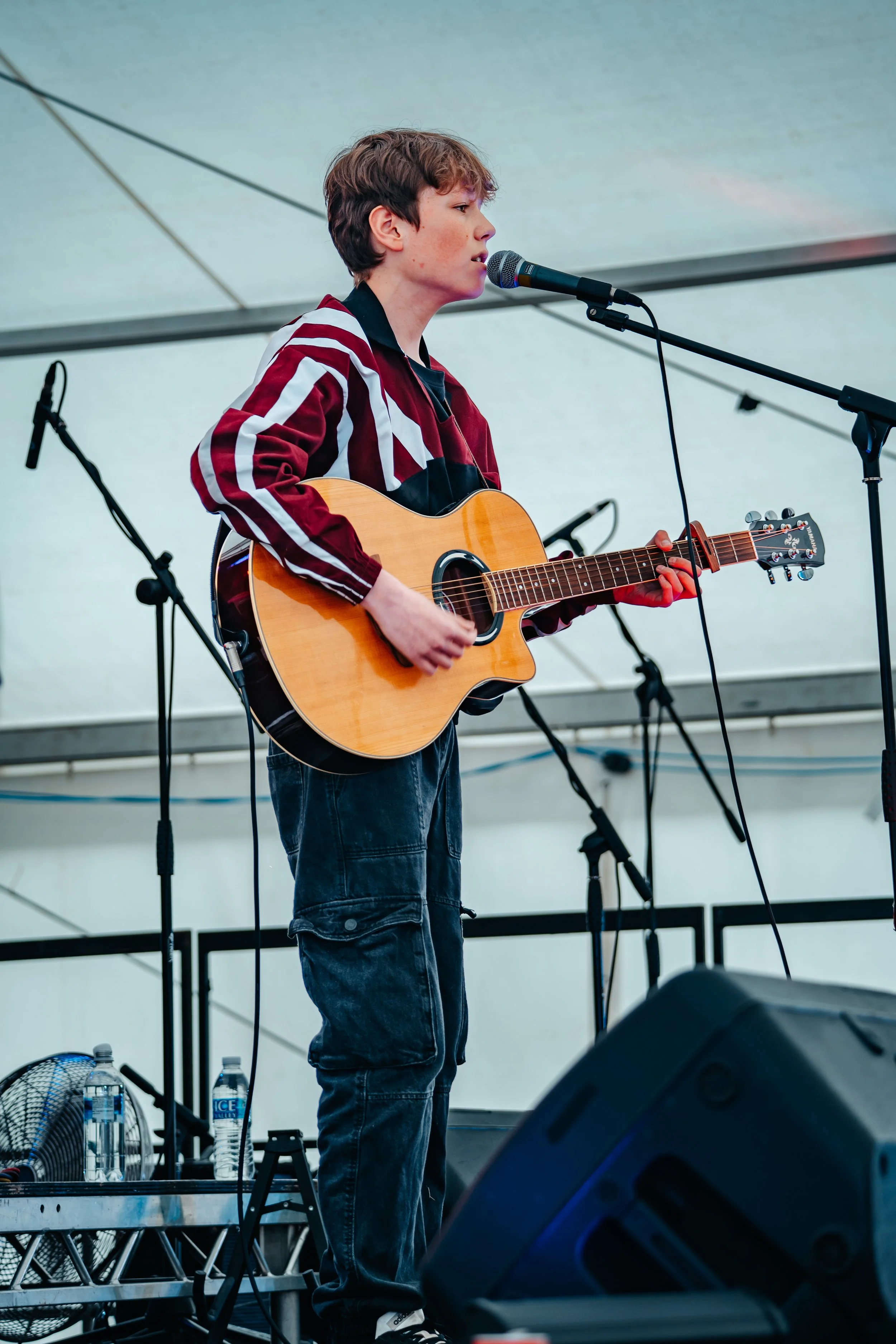 A young boy performing on stage with a guitar and microphone, under a tent, with sound equipment and water bottles nearby.