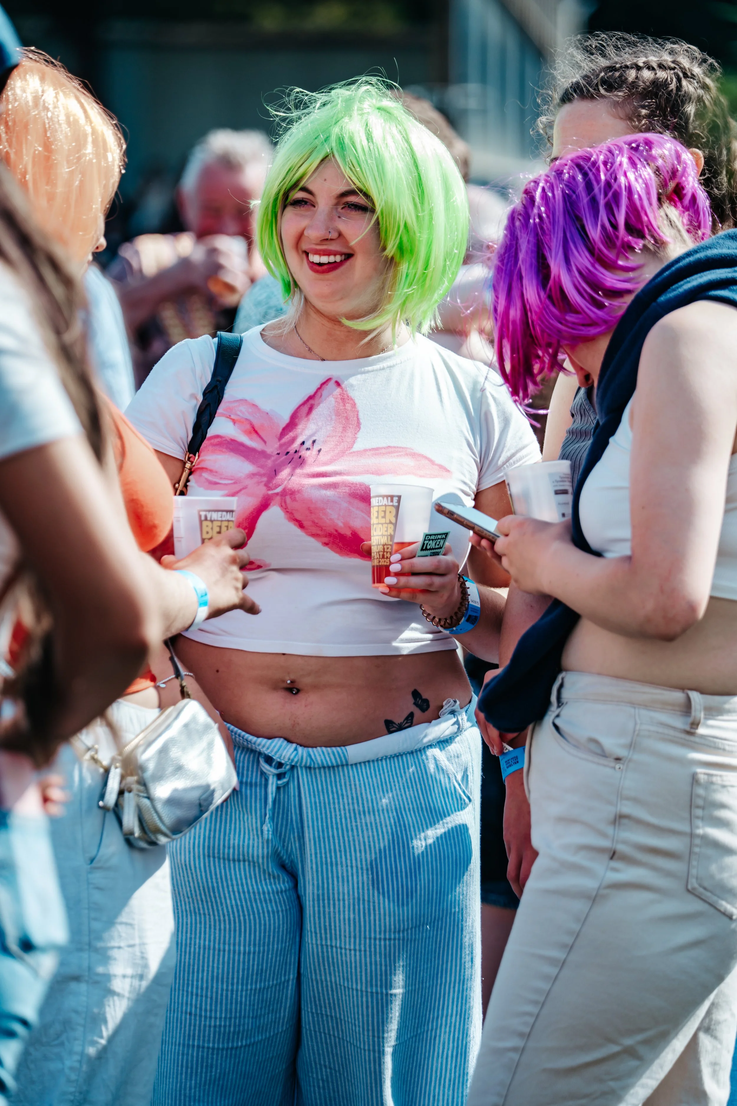 Group of young women at outdoor festival, colorful wigs, casual summer clothing, holding drinks, socializing.