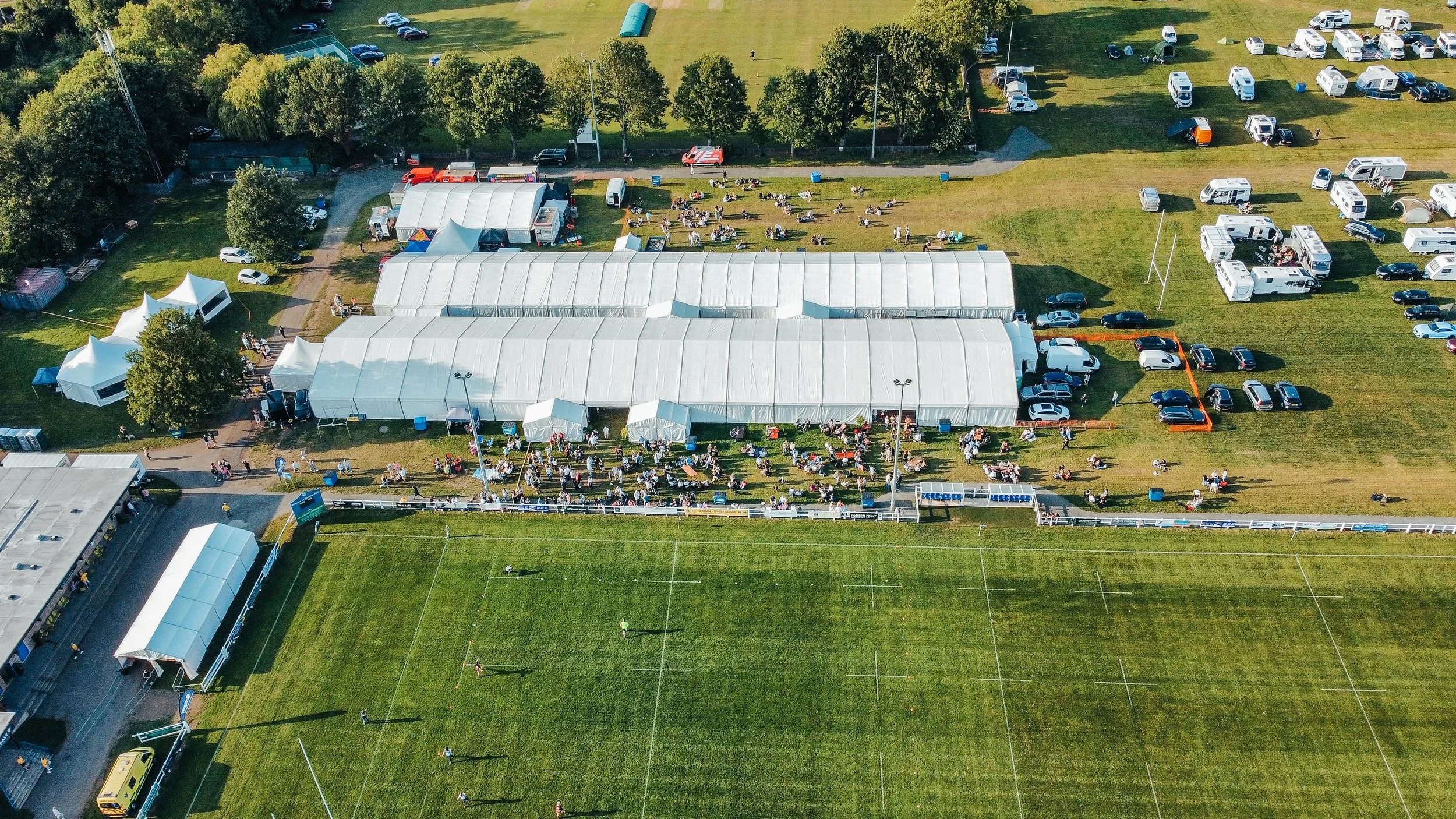 Aerial view of a sports event with tents, parking lot, and spectators on the grassy field and surrounding area.