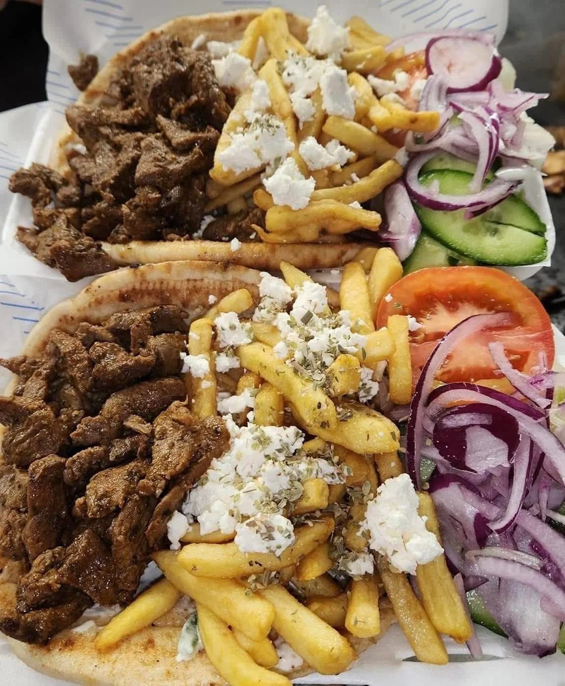 Two plates of Greek gyros with meat, French fries, feta cheese, herbs, tomato slices, cucumbers, and sliced onions.