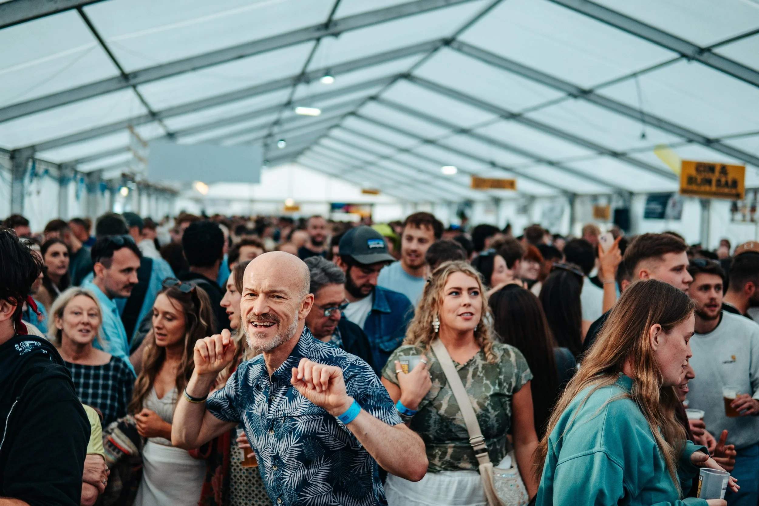 Crowd of people inside a large tent at an event or festival, with some individuals dancing and others holding drinks.