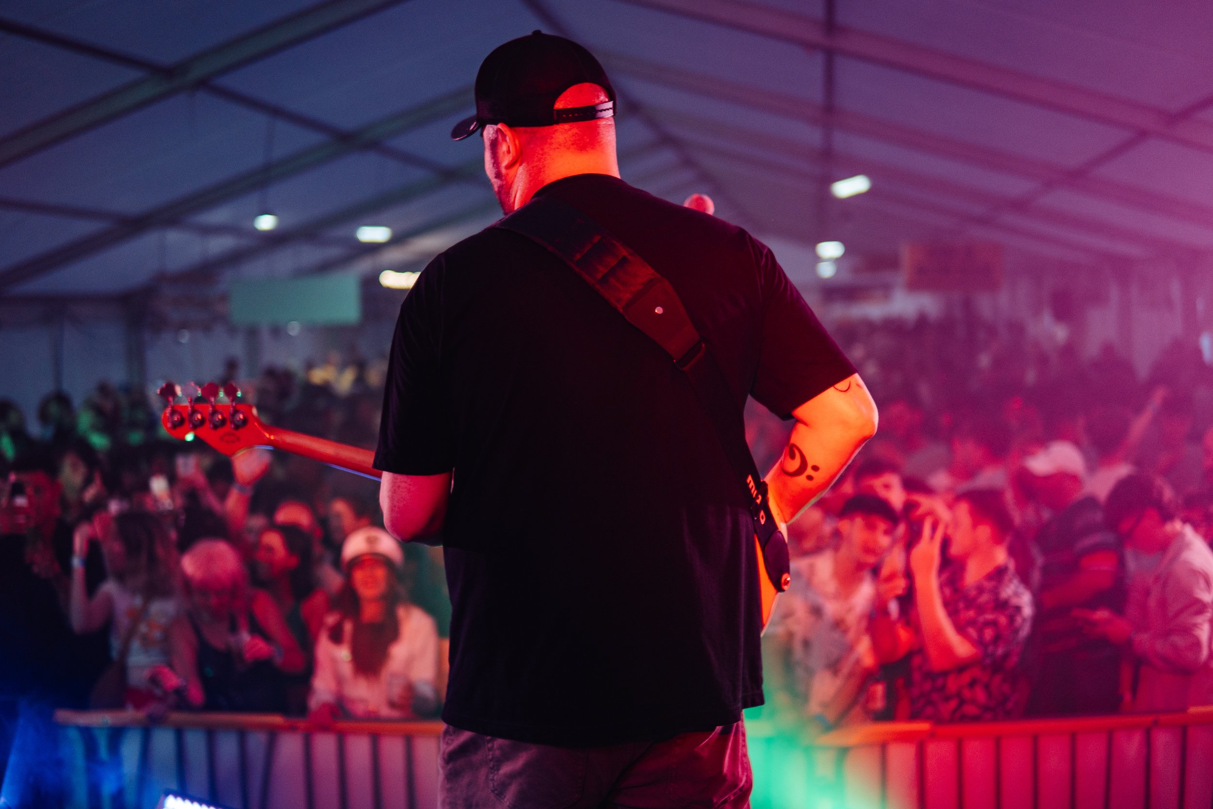 Back view of a guitarist performing on stage at a concert, with a crowded audience in the background under a large tent.