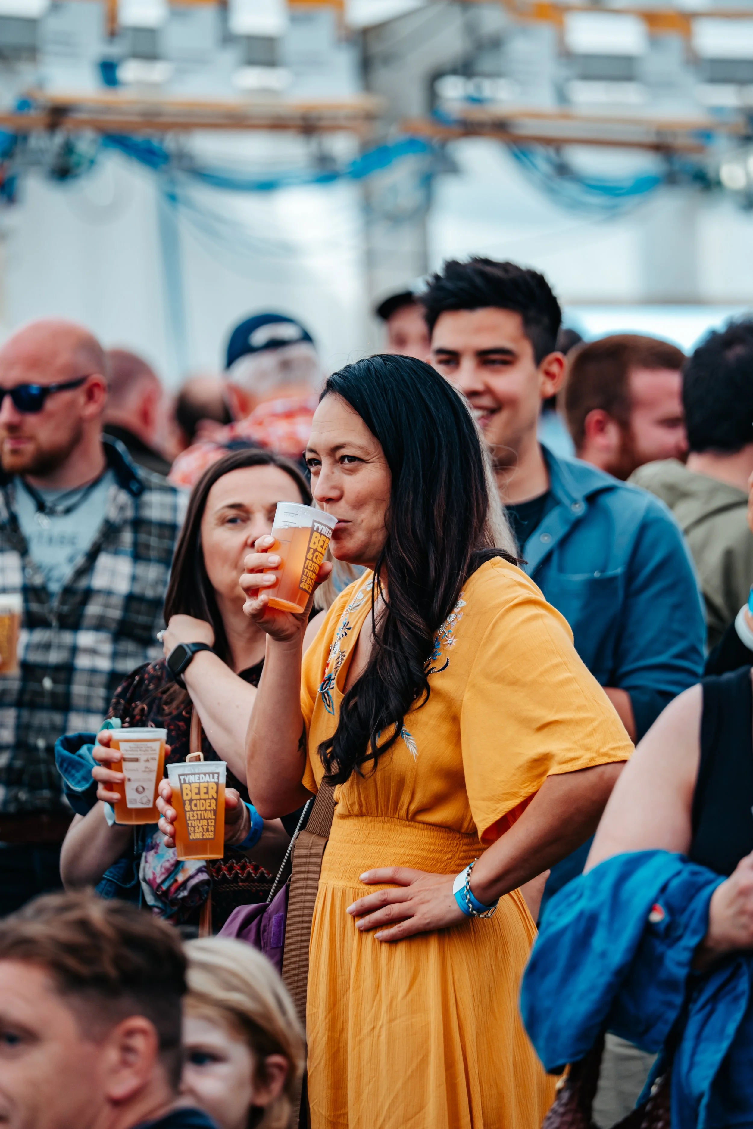 A woman in a yellow dress drinking beer at a crowded outdoor festival.