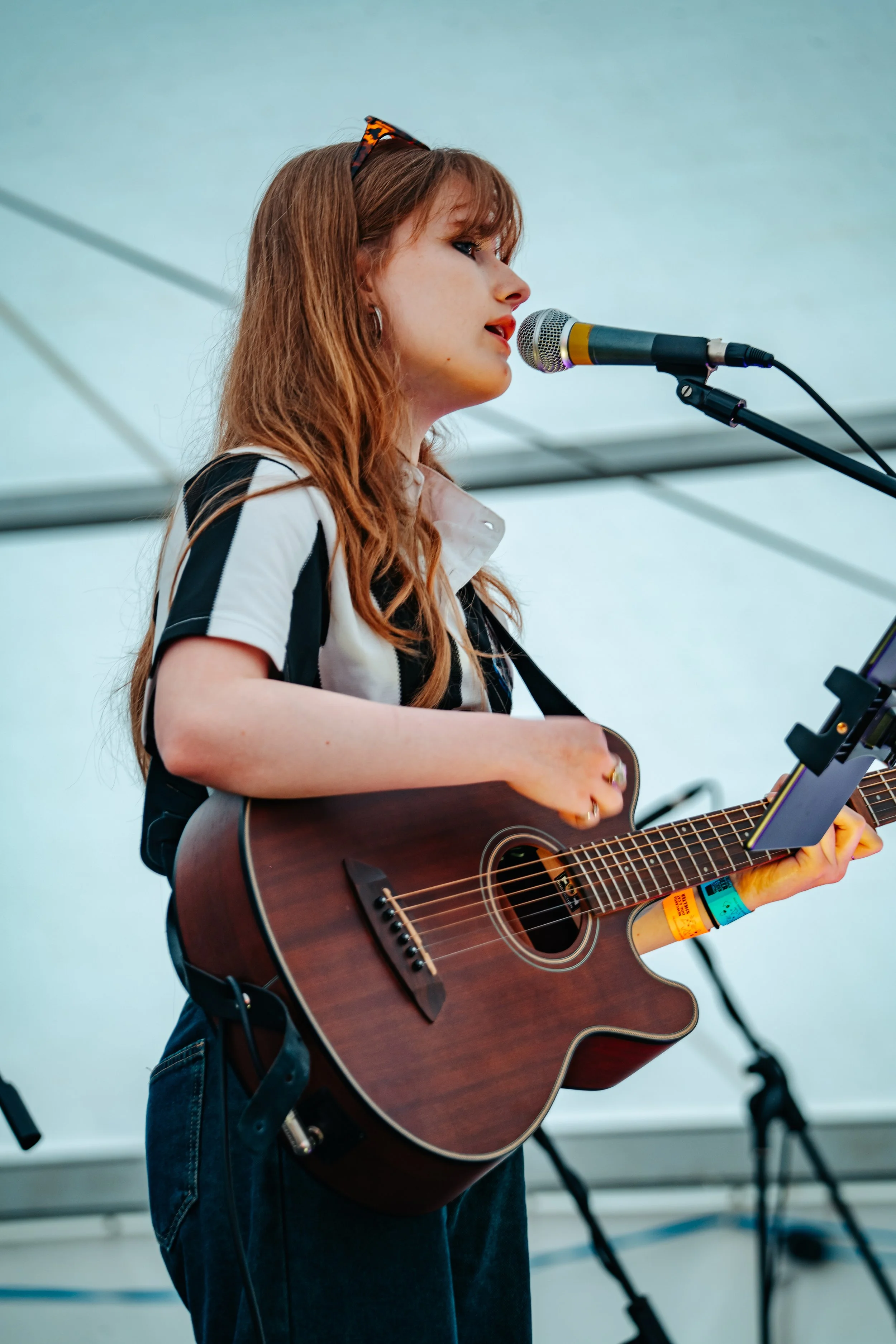 Young woman with red hair wearing sunglasses and a striped shirt, playing an acoustic guitar and singing into a microphone during a live performance.