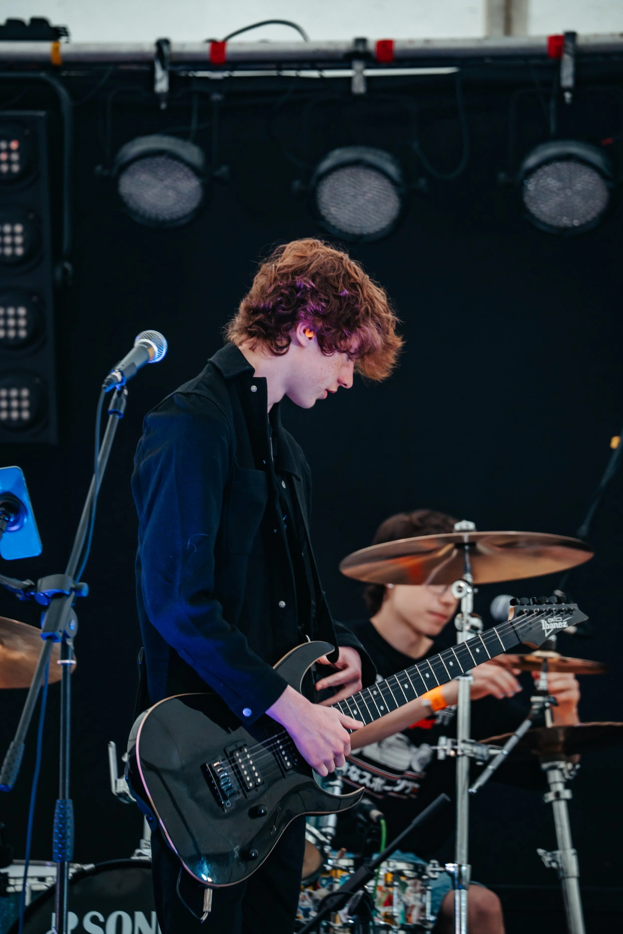 A young musician with curly hair playing an electric guitar on stage during a live performance, with another musician playing drums in the background.