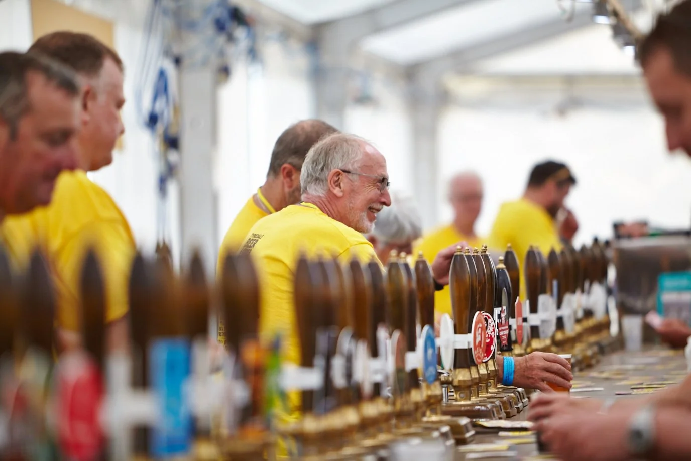 Older man with glasses and gray hair smiling, sitting at a bar with multiple beer taps in front of him, wearing a yellow shirt.