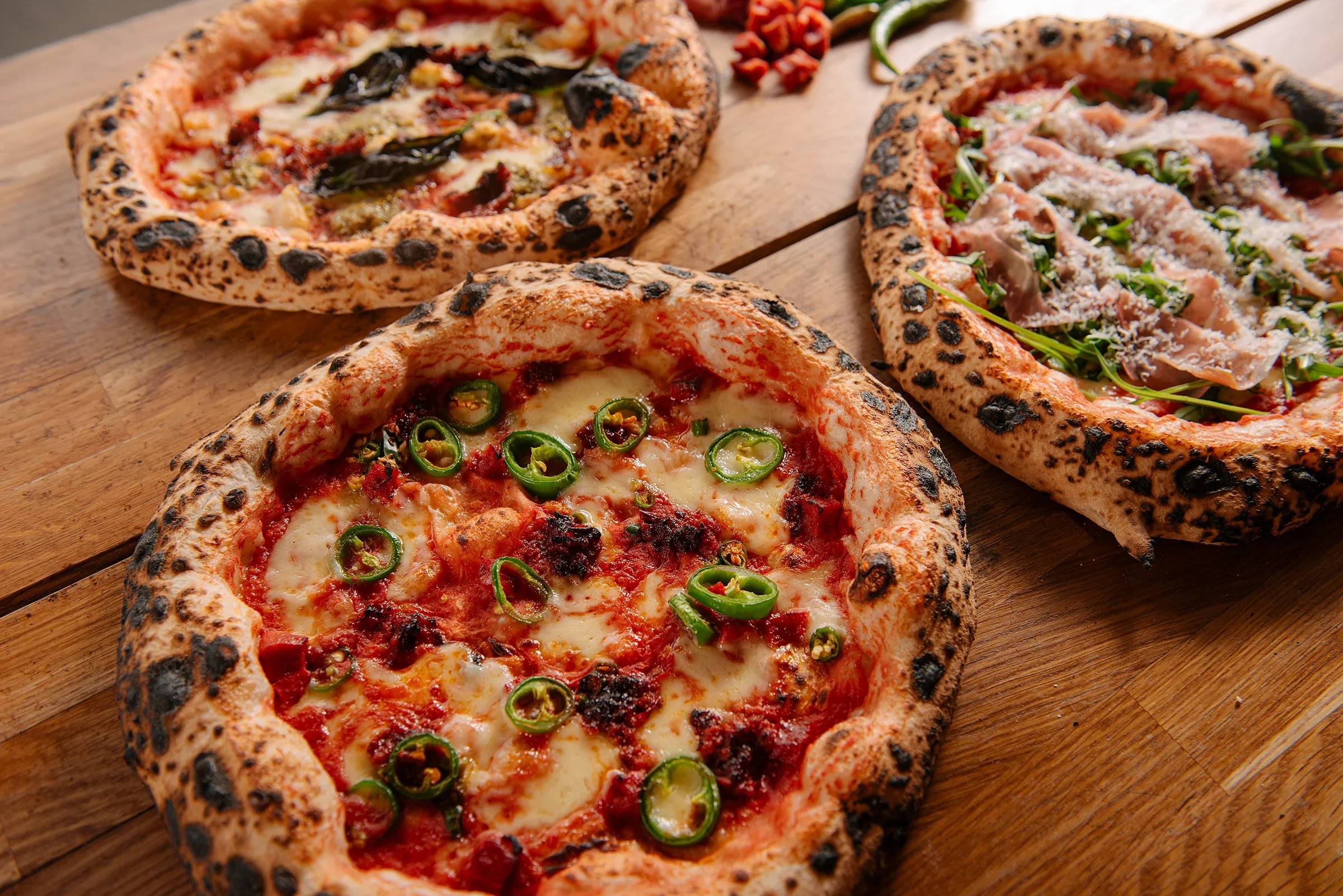 Three wood-fired pizzas on a wooden table, one with jalapeños and cheese, another with tomato sauce and various toppings, and the third topped with prosciutto, arugula, and cheese.