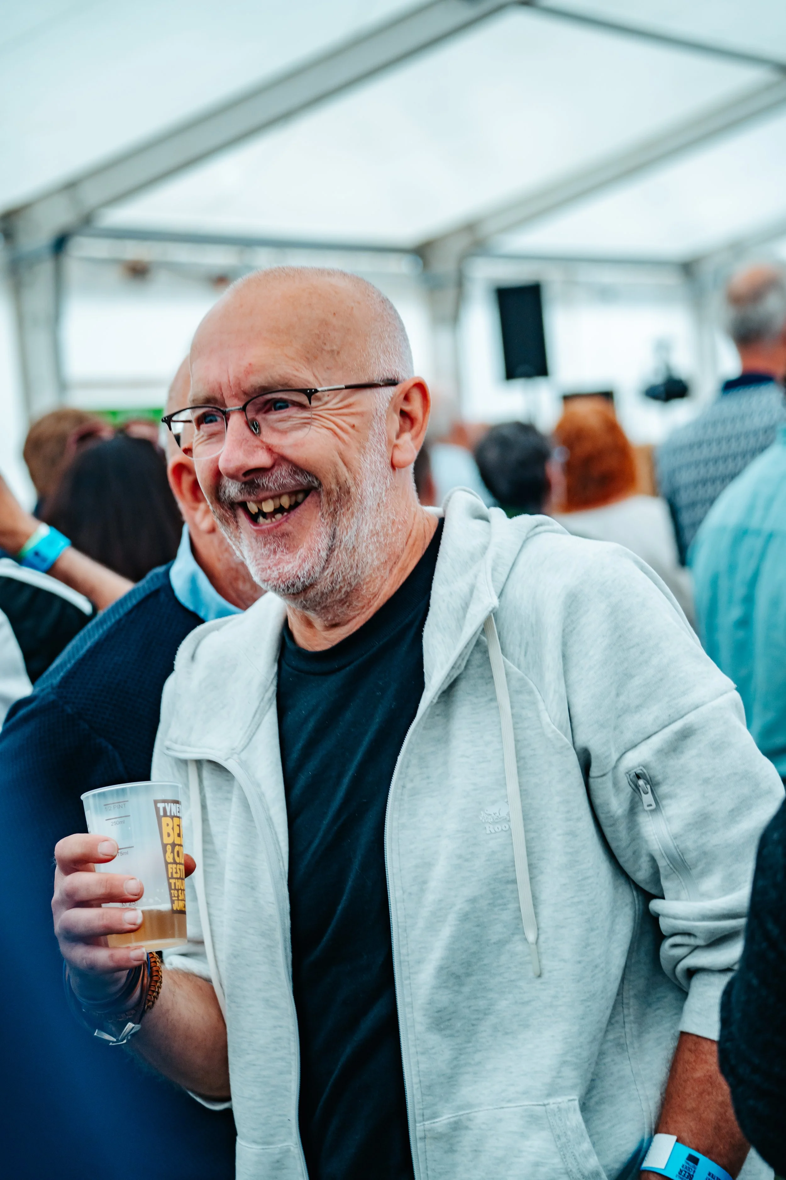 A smiling man with glasses, a bald head, and a gray beard, holding a plastic cup, at a crowded indoor event or festival.