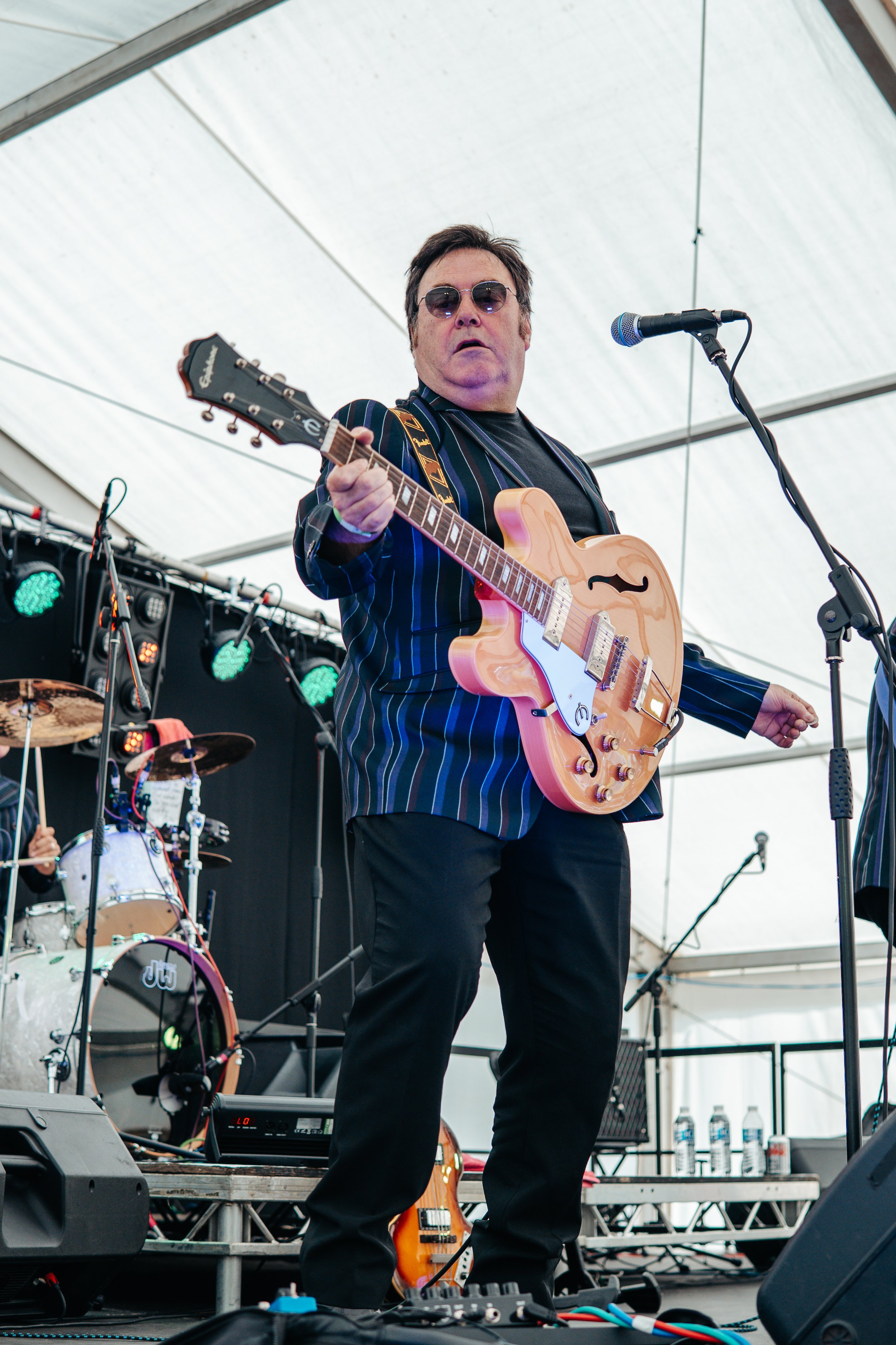 Male musician wearing sunglasses and a striped blazer playing an electric guitar on stage.