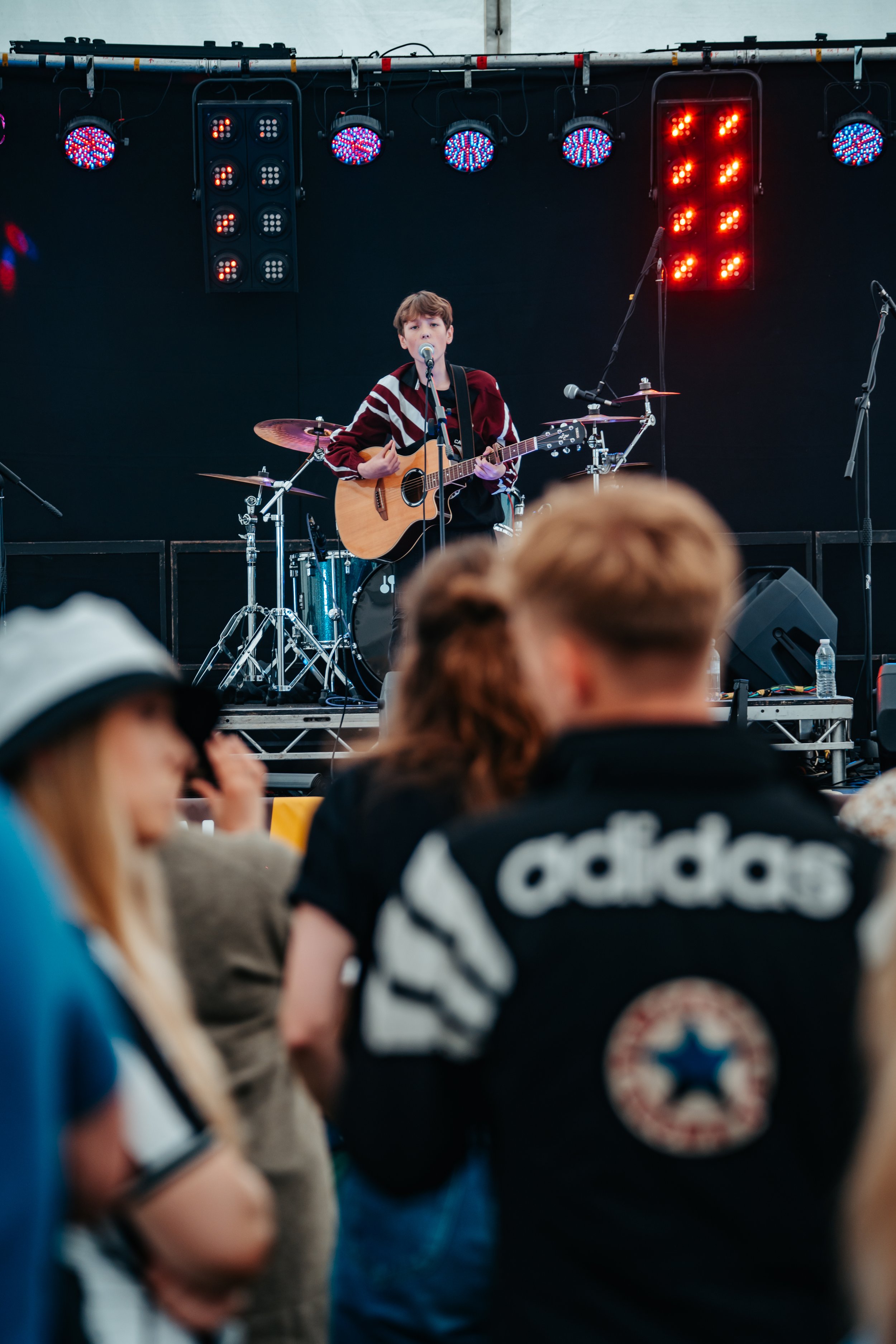 A young male musician performs on stage with an acoustic guitar and microphone, surrounded by a drum set and stage lights, with an audience watching.