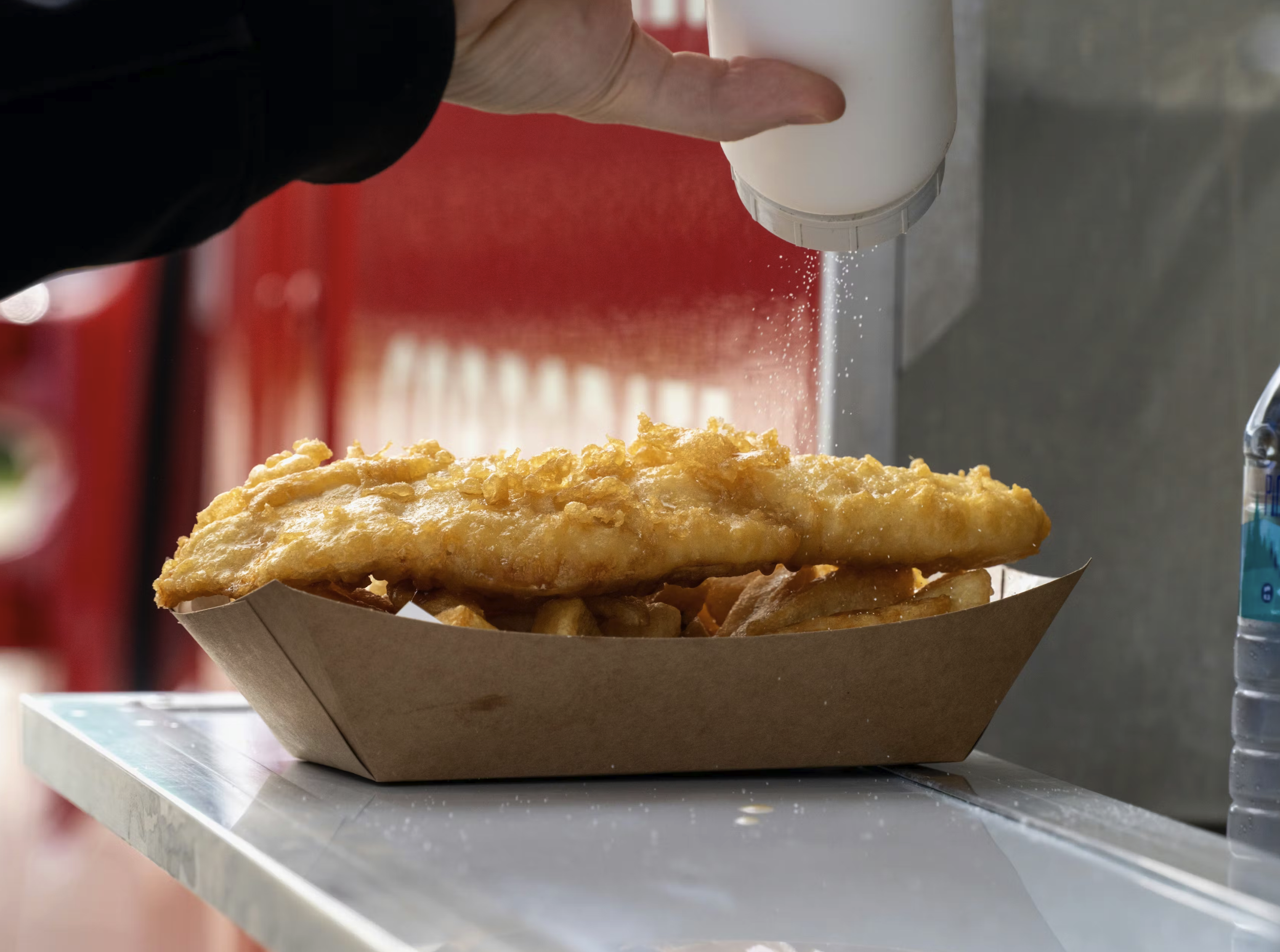 A hand sprinkling salt on a fried fish and chips in a paper tray on a table near a water bottle