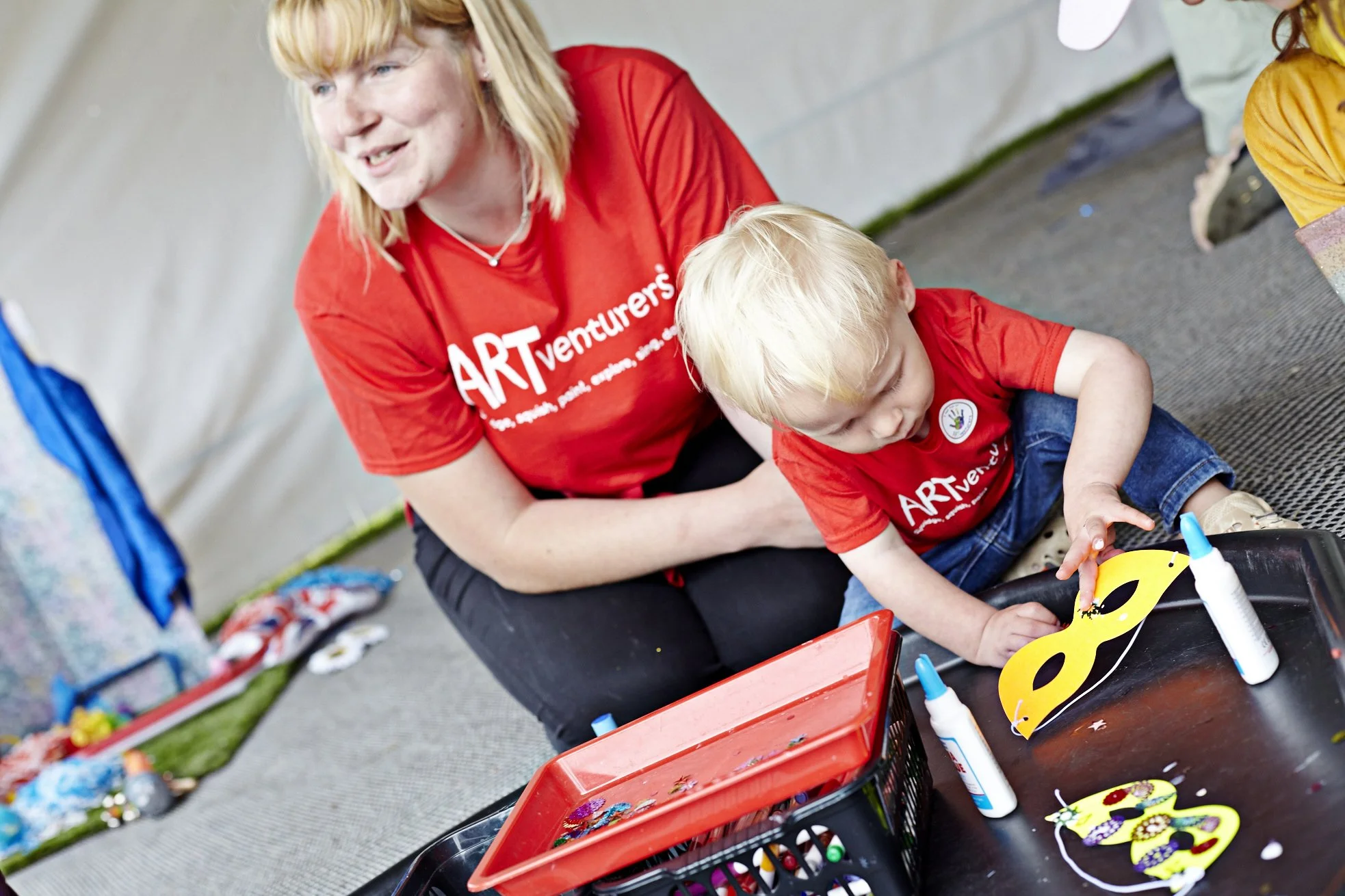 A woman and a young boy sit on the ground at an arts and crafts table, both wearing red 'ARTventures' shirts. The boy is focused on making a yellow paper mask with black eye holes, surrounded by glue bottles and scissors. Colorful materials and supplies are scattered around the table and on the background.